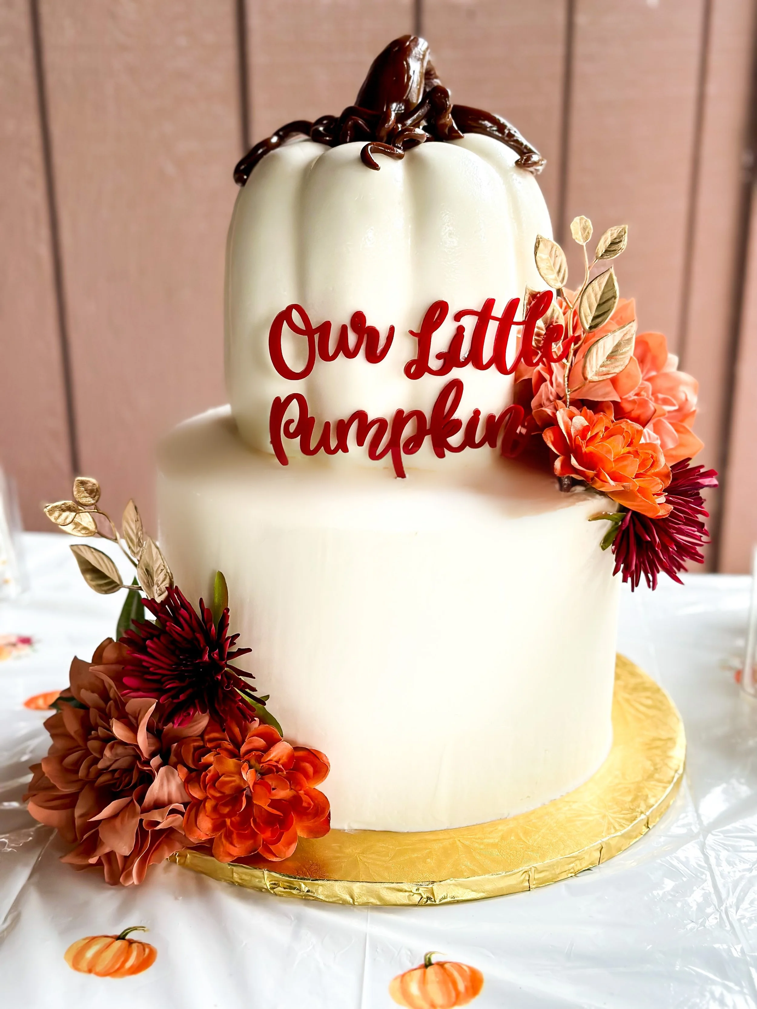 Two-tiered white cake with a chocolate spider on top, decorated with orange and burgundy flowers, and a red sign that reads "Our Little Pumpkin."