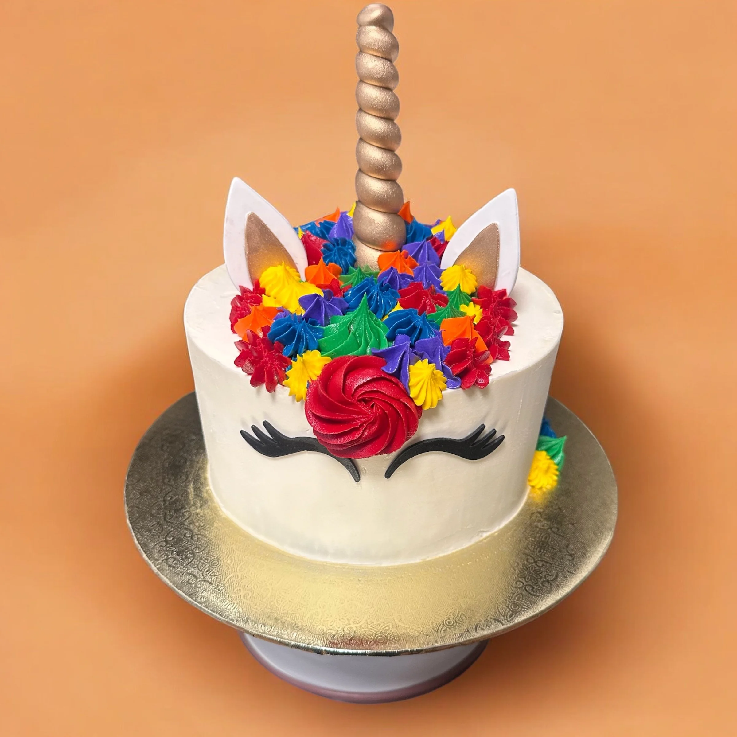 Unicorn-themed birthday cake with white frosting, rainbow-colored piped flowers, a red frosting swirl, a unicorn horn, white ears with gold accents, and closed eyes with long eyelashes, on a golden cake board.