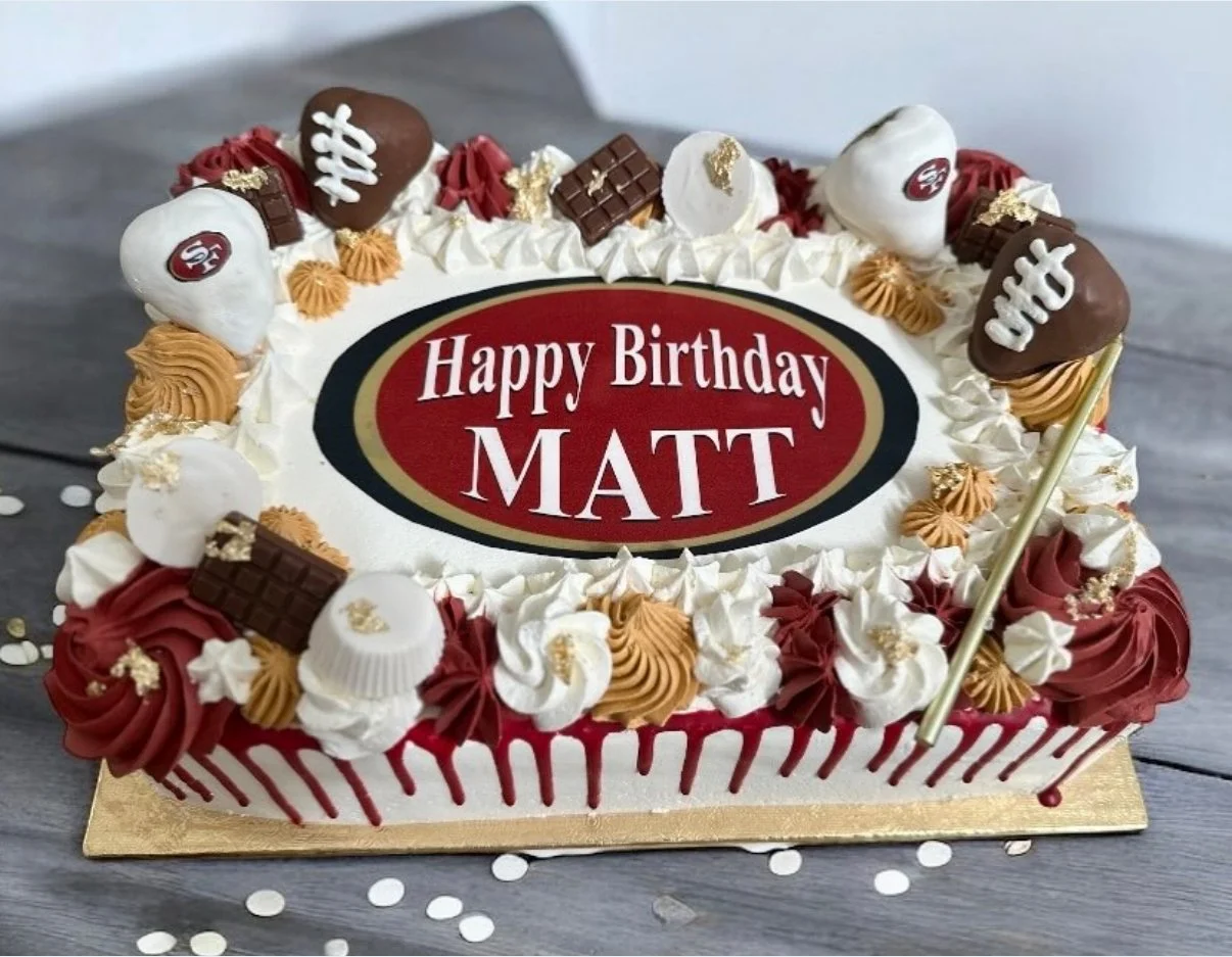 Birthday cake decorated with white, red, and brown frosting, chocolate, and gold accents, with a message "Happy Birthday MATT" written on an edible plaque.