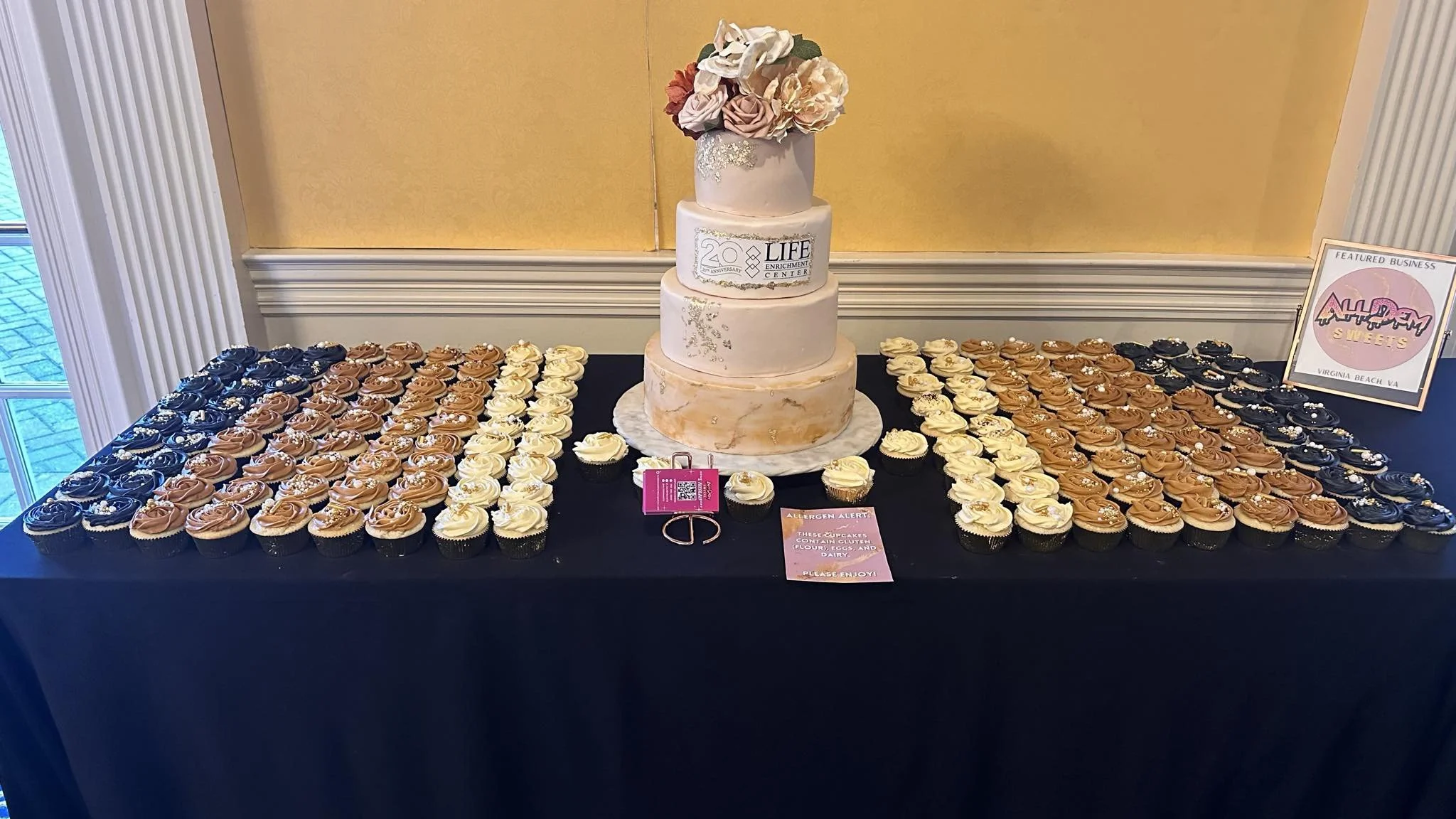 A celebration table with a four-tiered cake with a flower topper and an 'LIFE Enrichment Center' logo, surrounded by cupcakes with gold, white, and black frosting, pearl decorations, and a sign with an allergy warning.