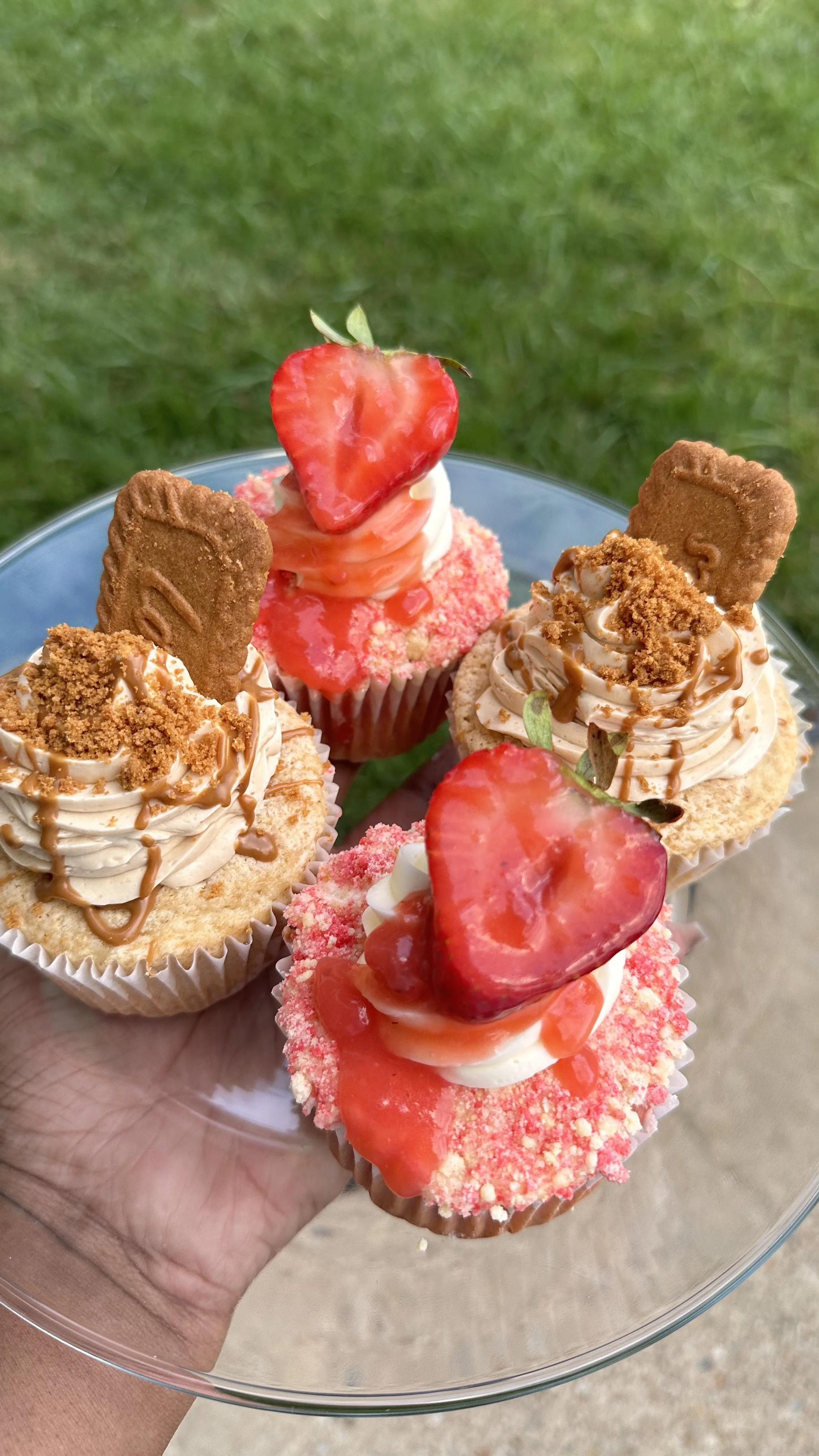 Three decorated cupcakes on a clear glass plate held outdoors with green grass background. Two cupcakes have strawberry slices on top, one with pink crumb coating, the others with swirl frosting, caramel drizzle, cookie crumbs, and cookie decorations