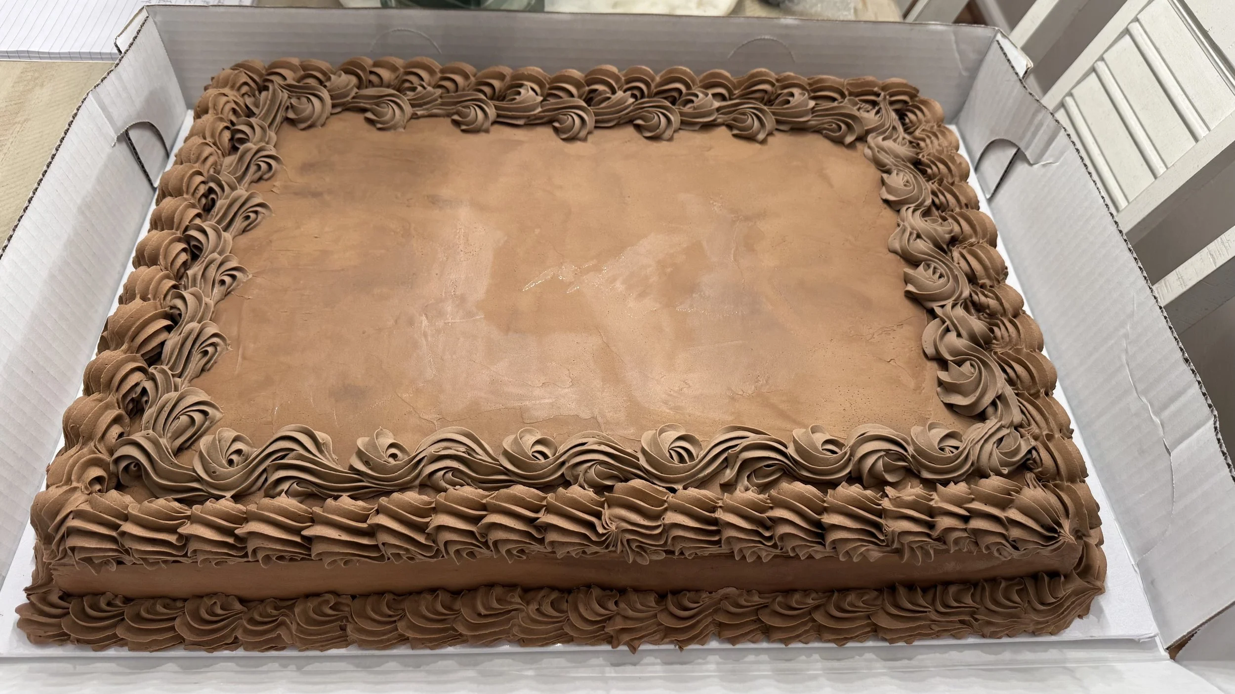 A large rectangular chocolate cake with piped chocolate frosting border sitting inside a white cardboard box.