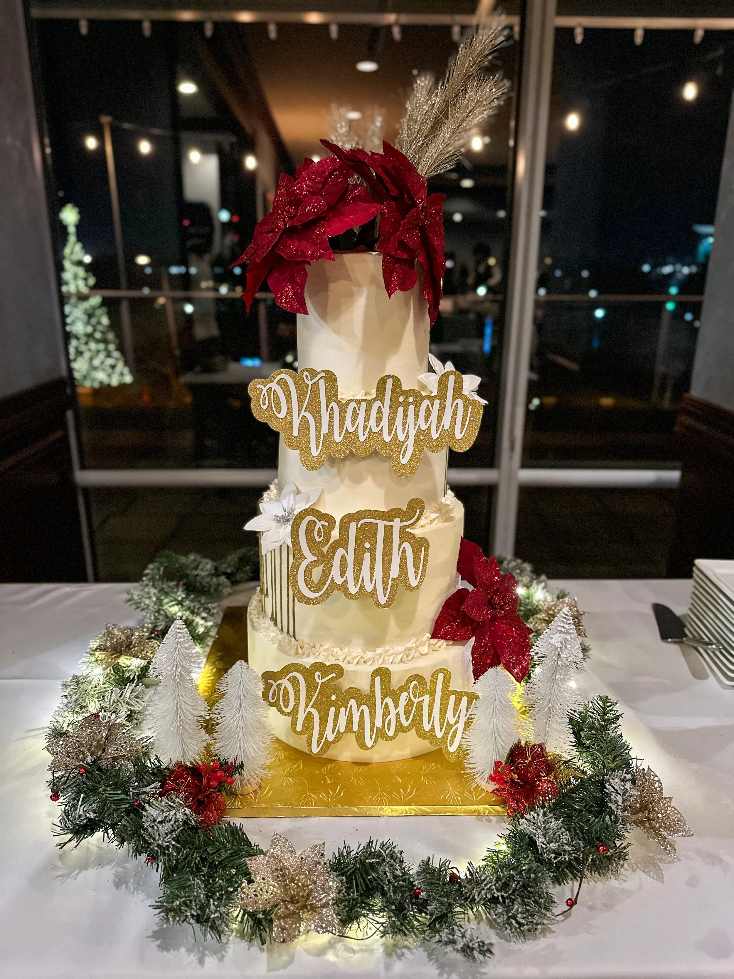 A tiered wedding cake decorated with red poinsettia flowers, white foliage, and gold names, surrounded by a Christmas-themed garland. The names on the cake are Khadijah, Edith, and Kimberly.