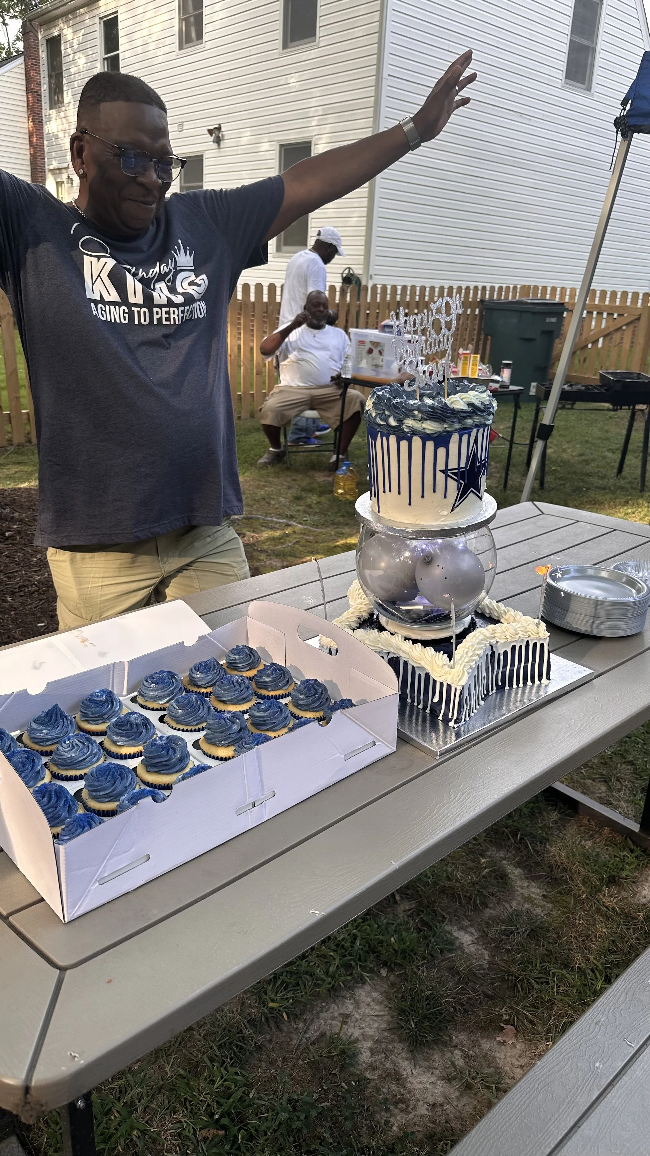 A man with glasses and a dark blue shirt with white lettering celebrating a birthday with a multiple-layer cake and cupcakes, outdoors near a house with a wooden fence, other people in the background, and a table with plates and decorations.