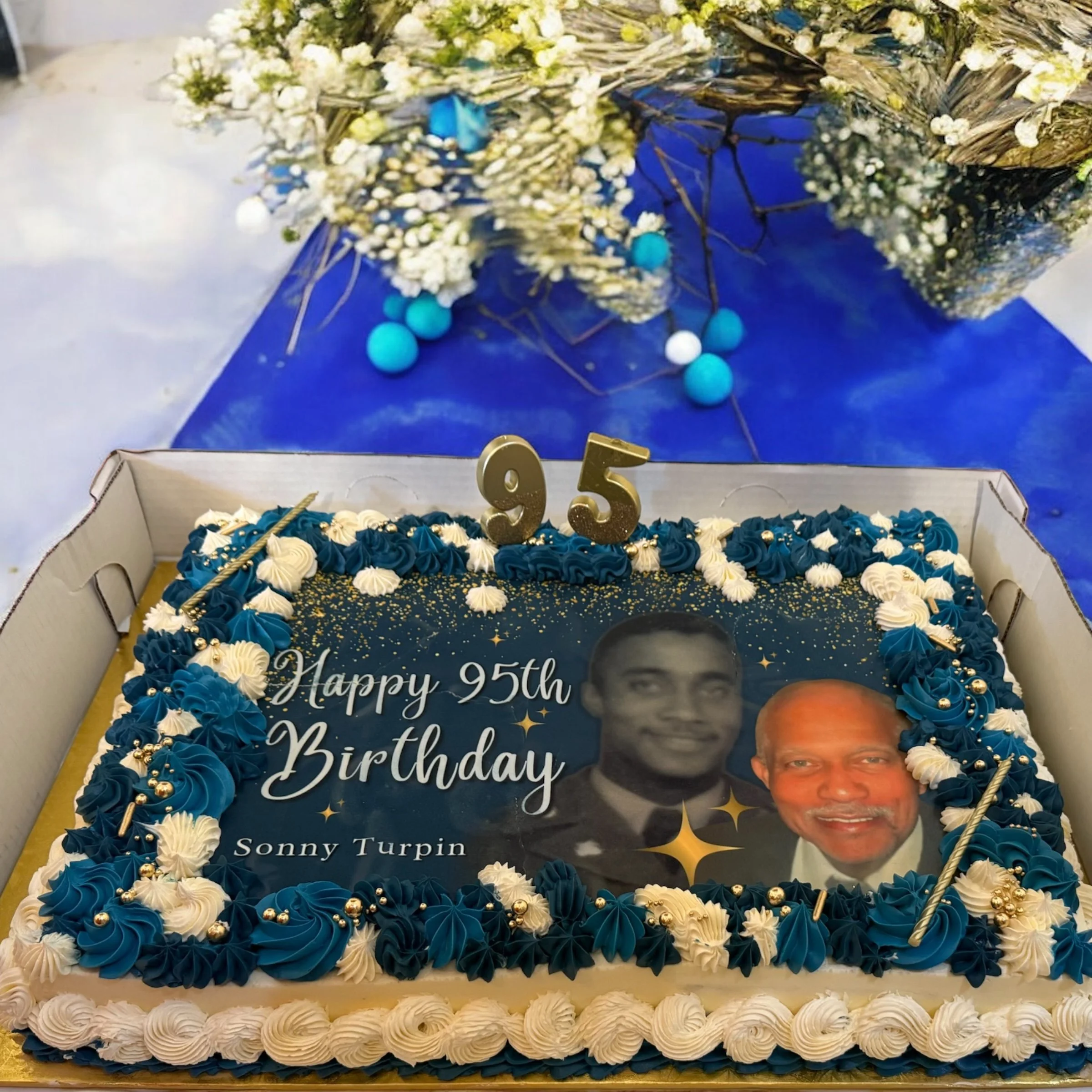 A birthday cake with blue and white icing, decorated with gold beads, featuring an edible photo of two men and the message "Happy 95th Birthday Sonny Turpin," topped with gold "95" candles, set on a table with a blue tablecloth and a flower arrangeme