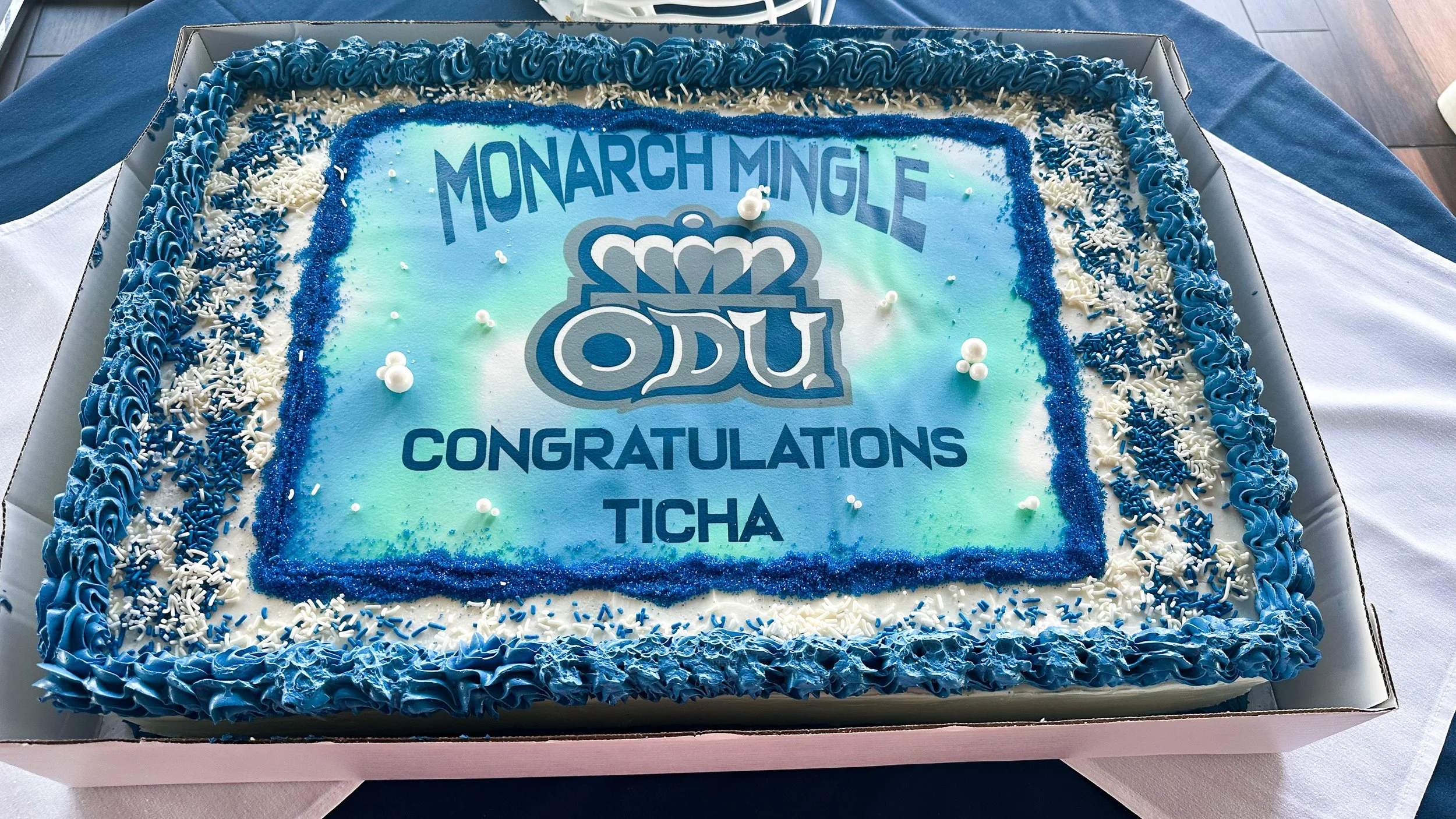 A rectangular cake with white frosting and blue decorative icing around the edges. The cake features a printed edible image with the Ohio Dominican University logo and the words 'Monarch Mingle Congratulations Ticha'.