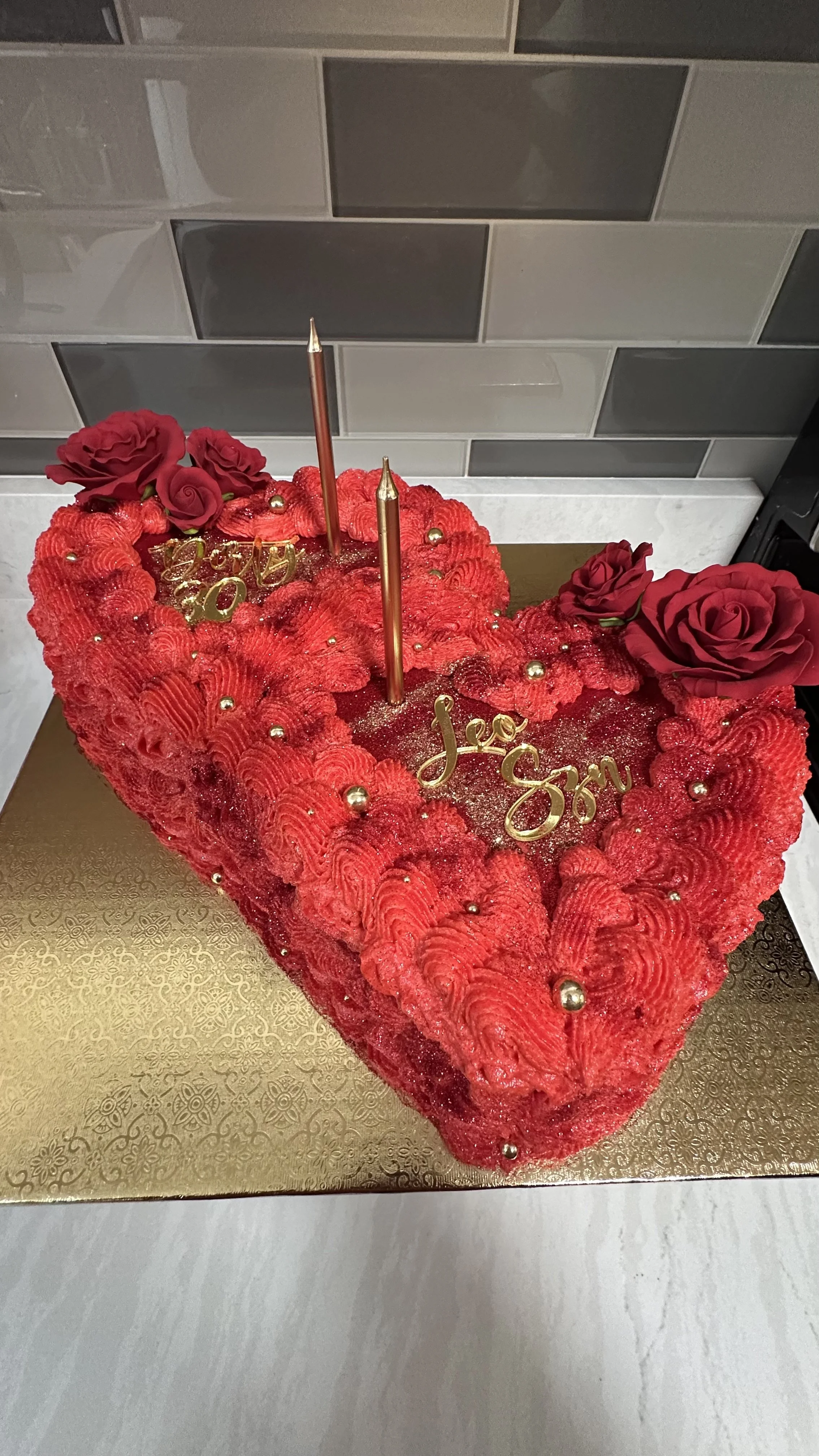 A heart-shaped red velvet cake decorated with red roses, gold ornaments, and two gold candles. The cake has gold 'Happy Birthday' and 'Love You' toppers and is placed on a gold cake board.
