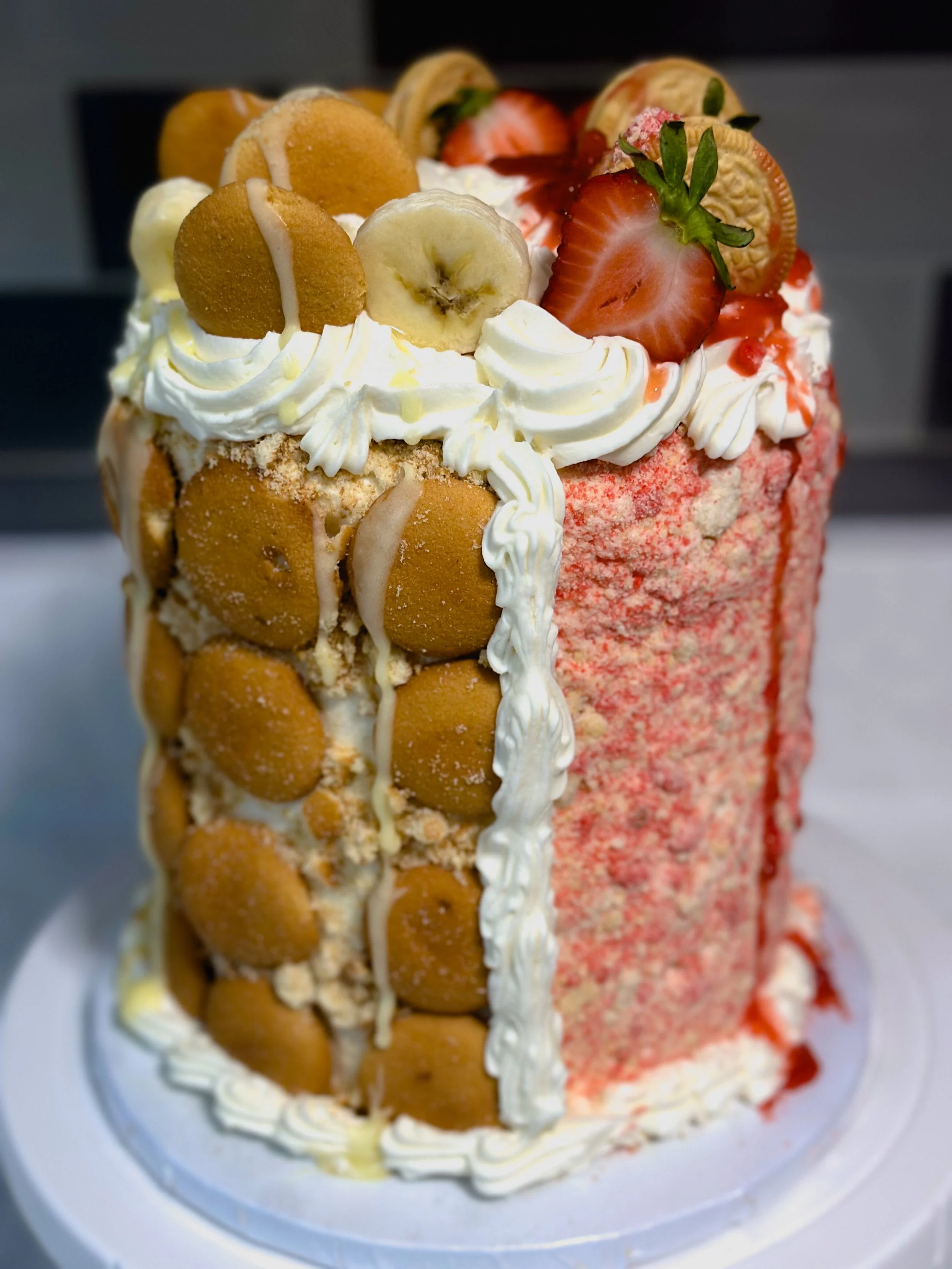 A tall ice cream cake decorated with whipped cream, strawberries, bananas, and small cookies on top. The sides are lined with mini cookies and white icing, with strawberry syrup drizzled over parts of the cake.