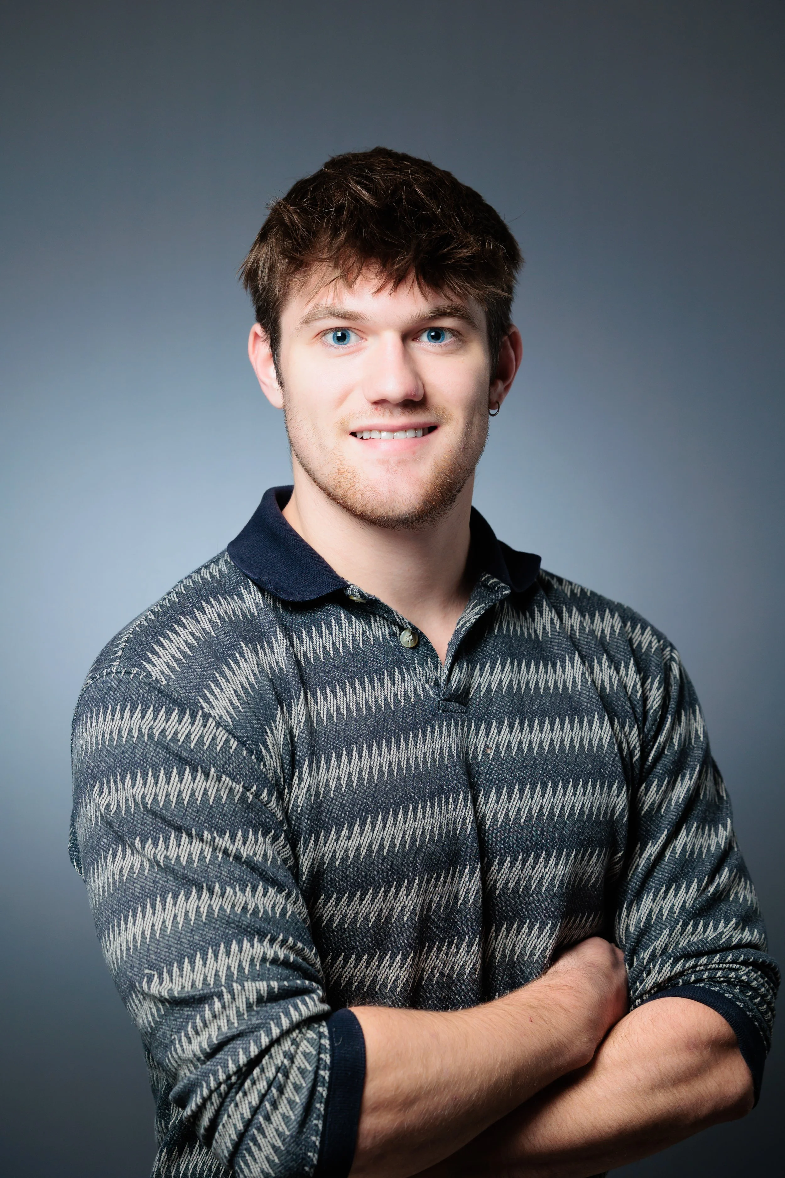 Portrait of a young man with brown hair and blue eyes, wearing a patterned dark sweater with a collar, crossing his arms in front of a plain gray background.