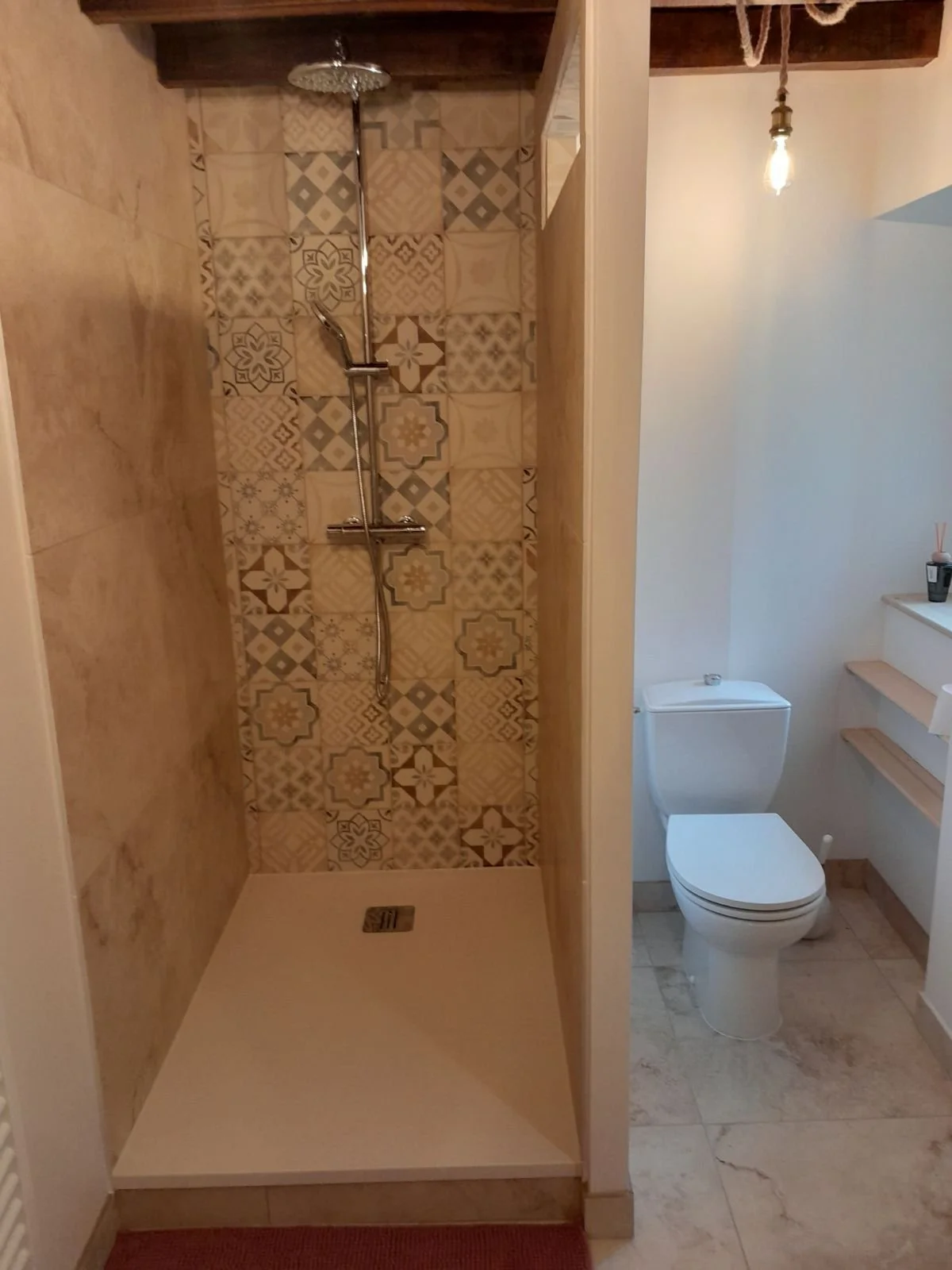 Salle de bain avec douche séparée, carrelage décoratif au mur, toilettes à droite, étagères en bois à côté des toilettes, plafond en bois et une lumière suspendue.