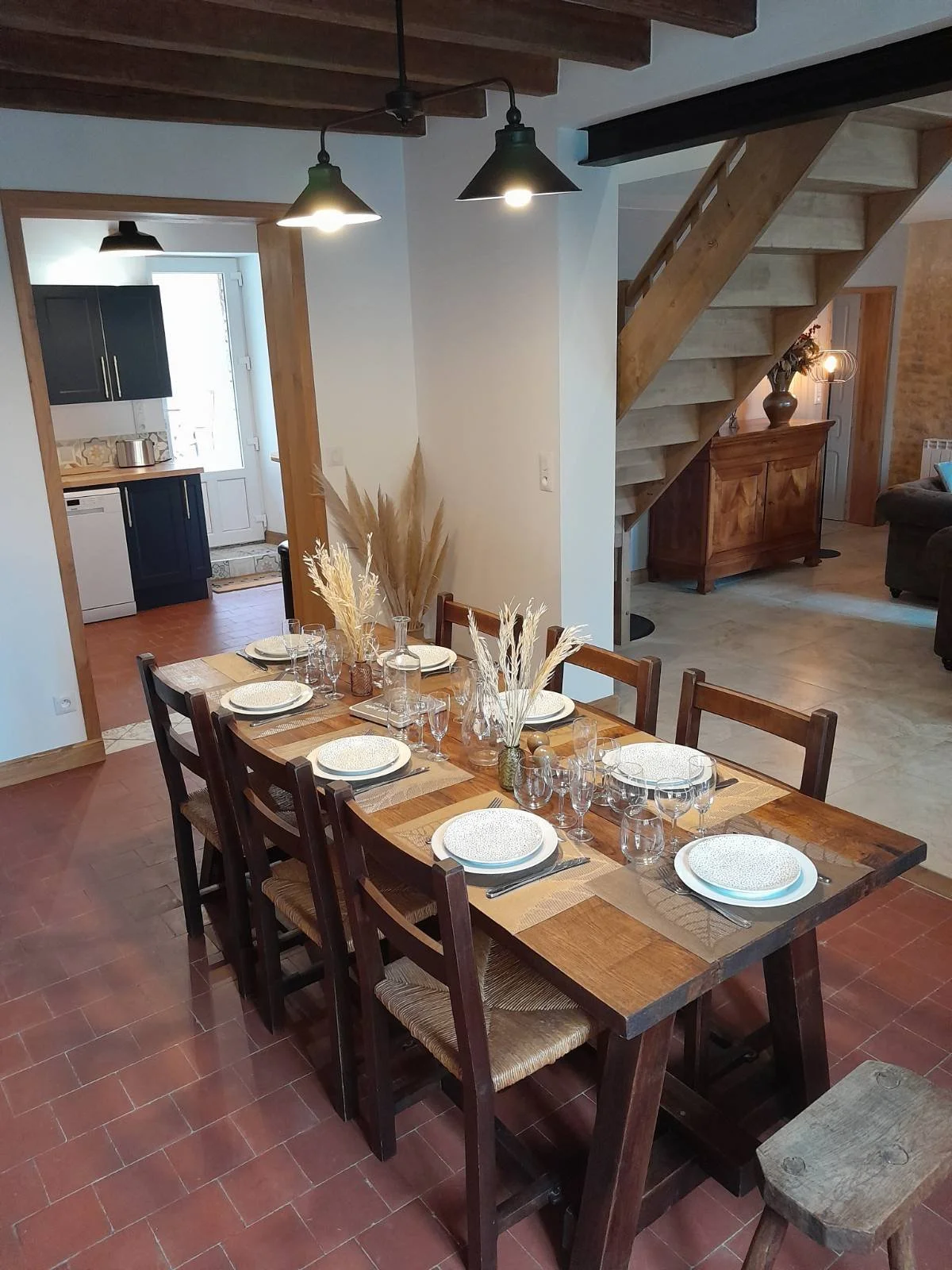 Salle à manger avec une grande table en bois, décorée de nappes, de vaisselle et de bouquets de plantes sèches. En arrière-plan, un escalier en bois mène à l'étage, et une porte fenêtre donne accès à la cuisine.