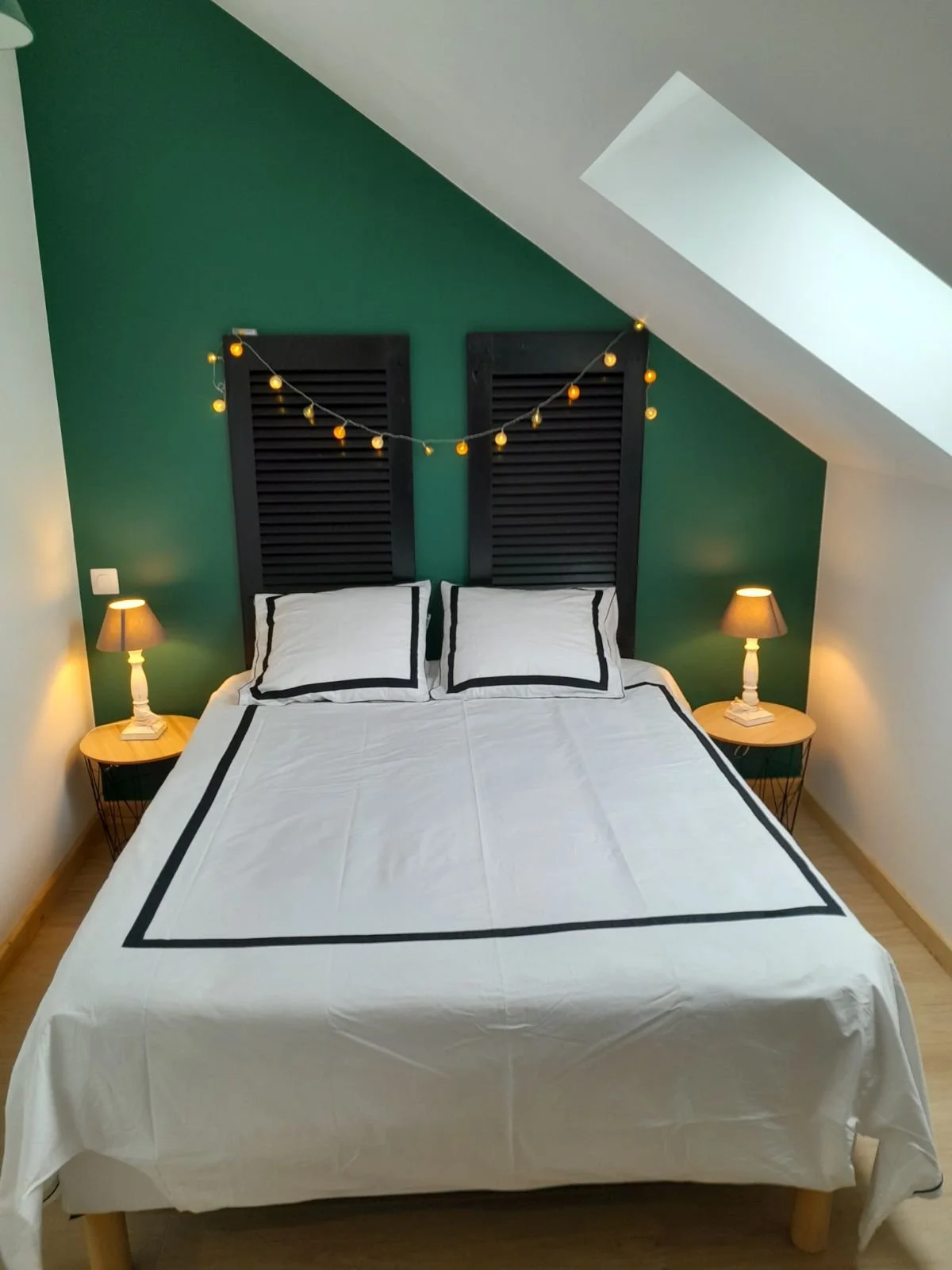 Lit simple avec oreillers, chevets avec lampes de chevet, guirlande lumineuse, mur vert derrière, plafond avec fenêtre de toit.