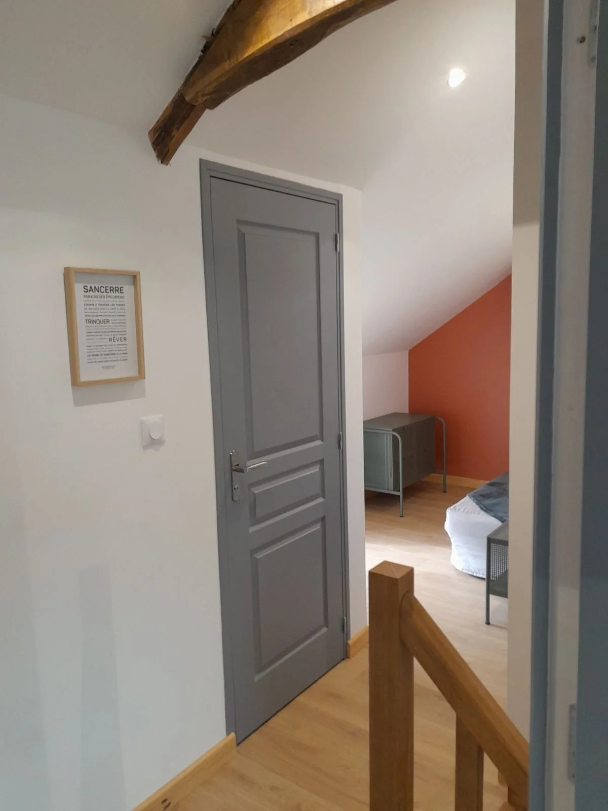 Une pièce d'intérieur avec un mur blanc, une porte grise, un sol en bois clair, un meuble en métal gris, un lit avec une couverture gris foncé, et un mur orange. Il y a une poutre en bois au plafond.