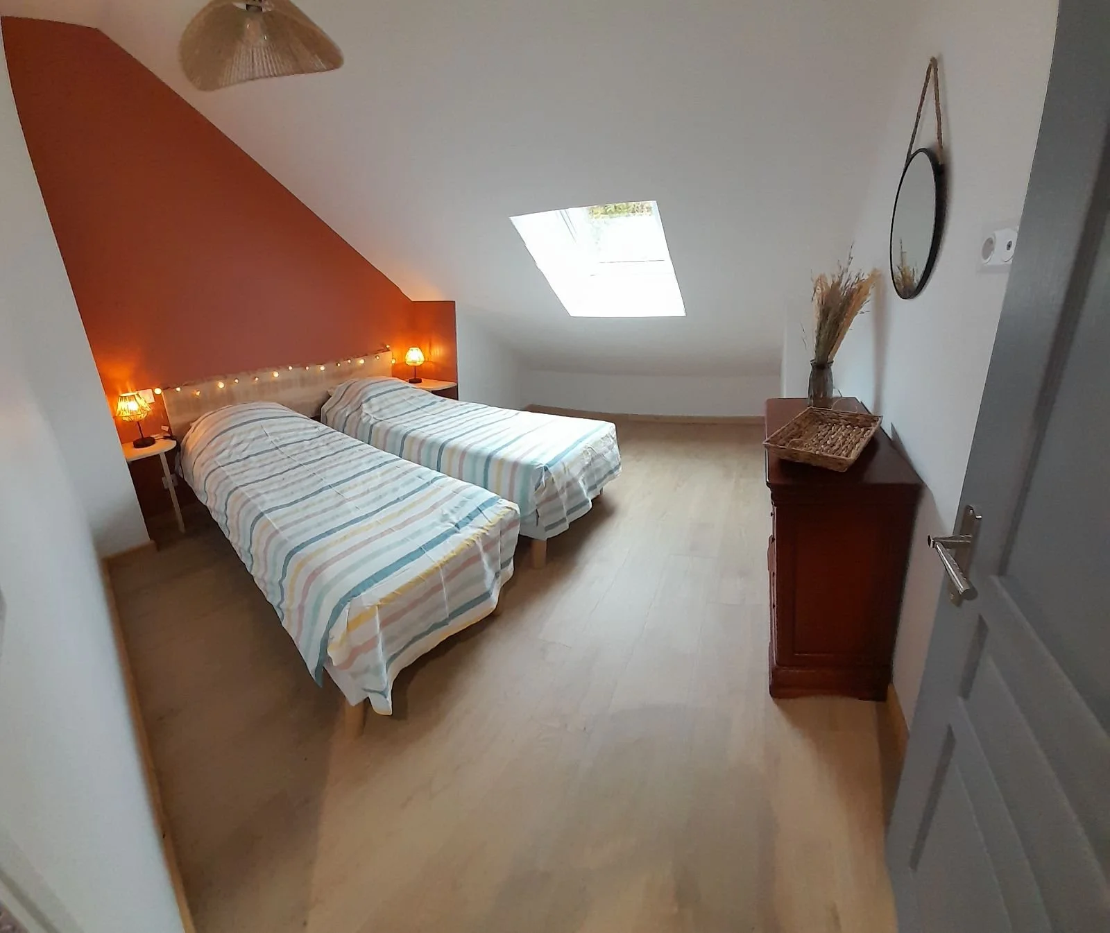 Chambre avec deux lits, tête de lit éclairée par deux petites lampes, mur rouge derrière les lits, fenêtre de toit, meuble en bois avec décoration, panier en osier.