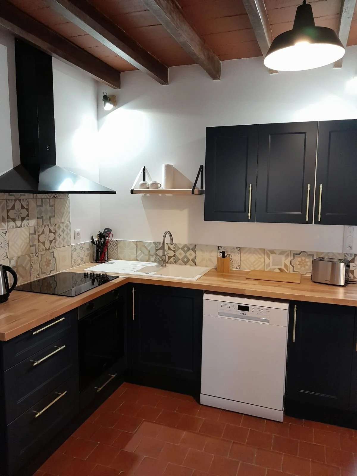 Cuisine moderne avec armoires noires, plan de travail en bois, crédence en carreaux décoratifs, évier sous la fenêtre, cuisinière électrique, hotte en acier au-dessus, petit électroménager en inox, lampe suspendue noire, mur blanc, sol en carreaux ro