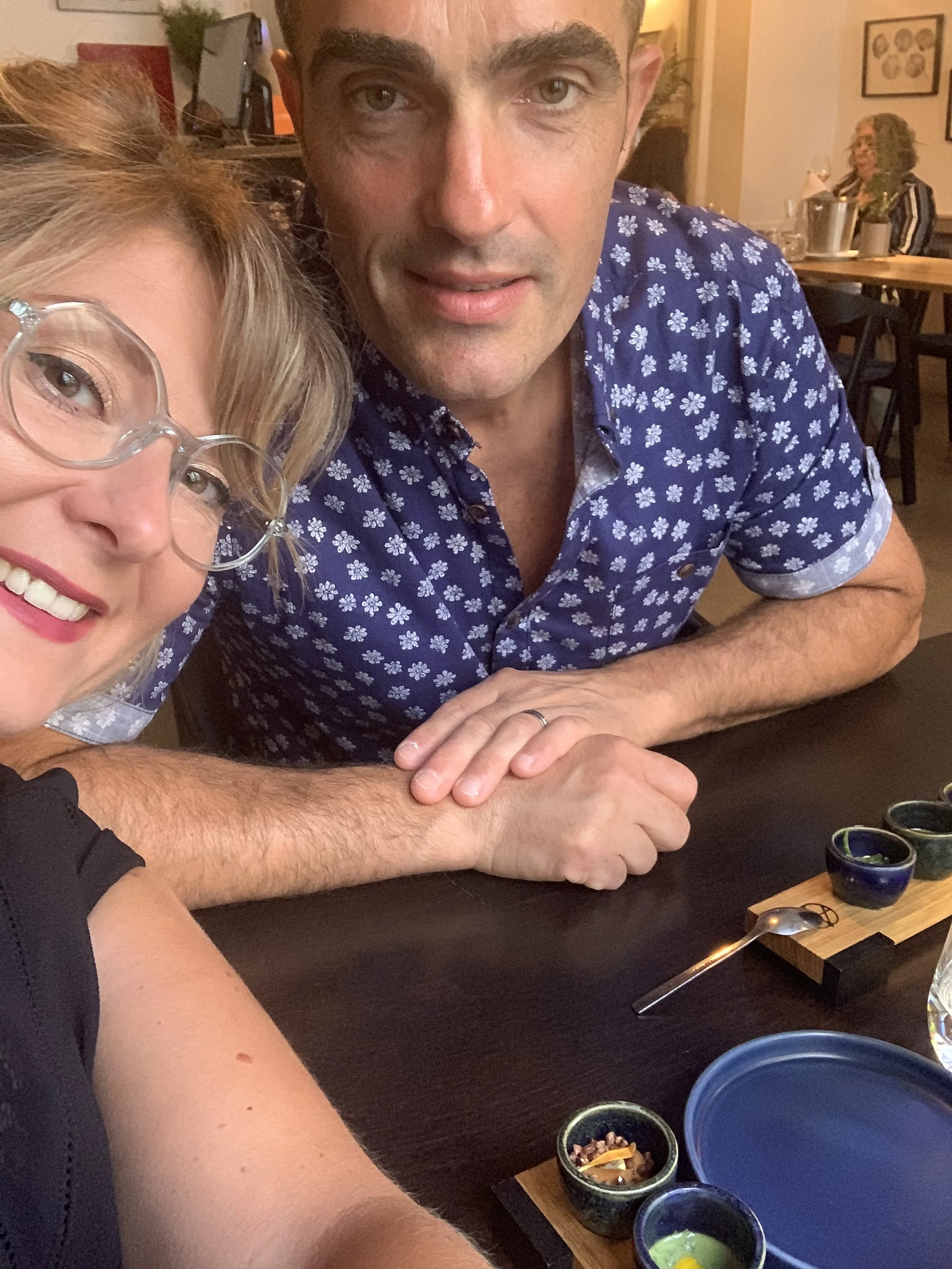 Deux personnes prennent un selfie dans un restaurant. La femme à gauche porte des lunettes transparentes et sourit, le homme à droite porte une chemise en coton à motifs de petites fleurs blanches et regarde l'appareil. Ils semblent être assis à une table avec des petits bols de sauces ou d'épices, et une planche en bois avec une cuillère en métal.