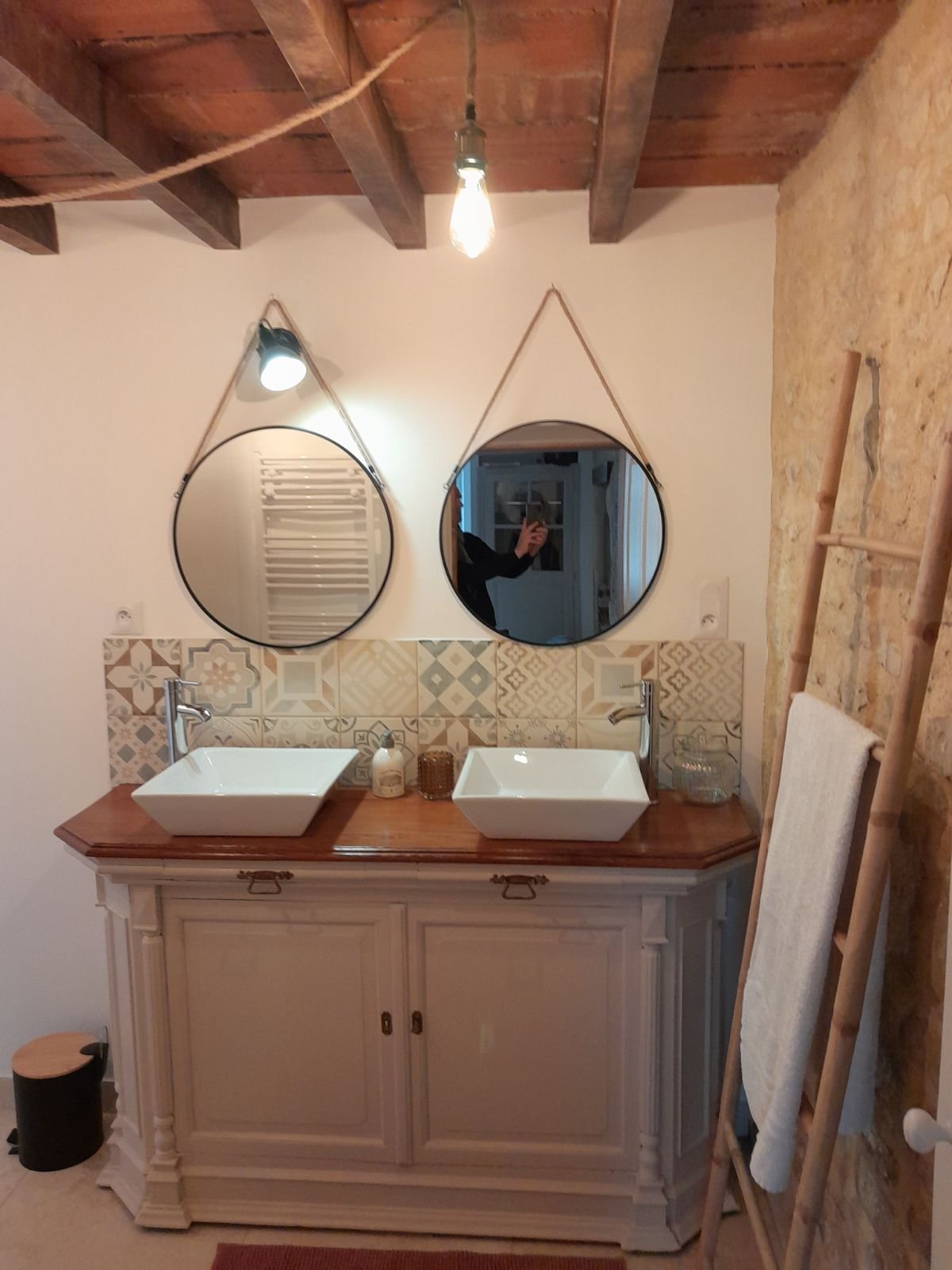 Sdb vintage avec double lavabo, deux miroirs ronds suspendus, un miroir à lampe, une desserte en bois blanche, un mur en pierre, un porte-serviettes en bois avec une serviette, un crochet noir pour brosse à dents, un panier en verre, un rouleau de pa