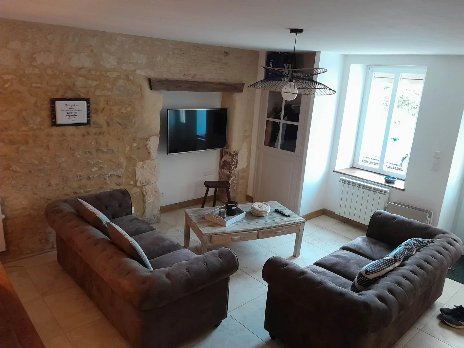 Salon avec deux canapés en velours gris, une table en bois au centre, un mur en pierre, une télévision fixée au mur, une fenêtre laissant entrer la lumière naturelle, un luminaire suspendu en forme de fleur.