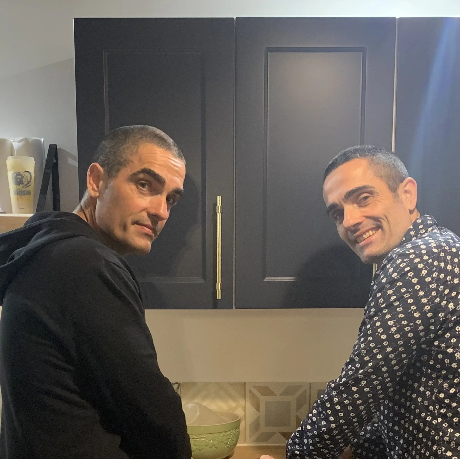 Deux hommes souriants qui se tiennent dans une cuisine, en face d'une armoire noire avec des poignées en métal doré.