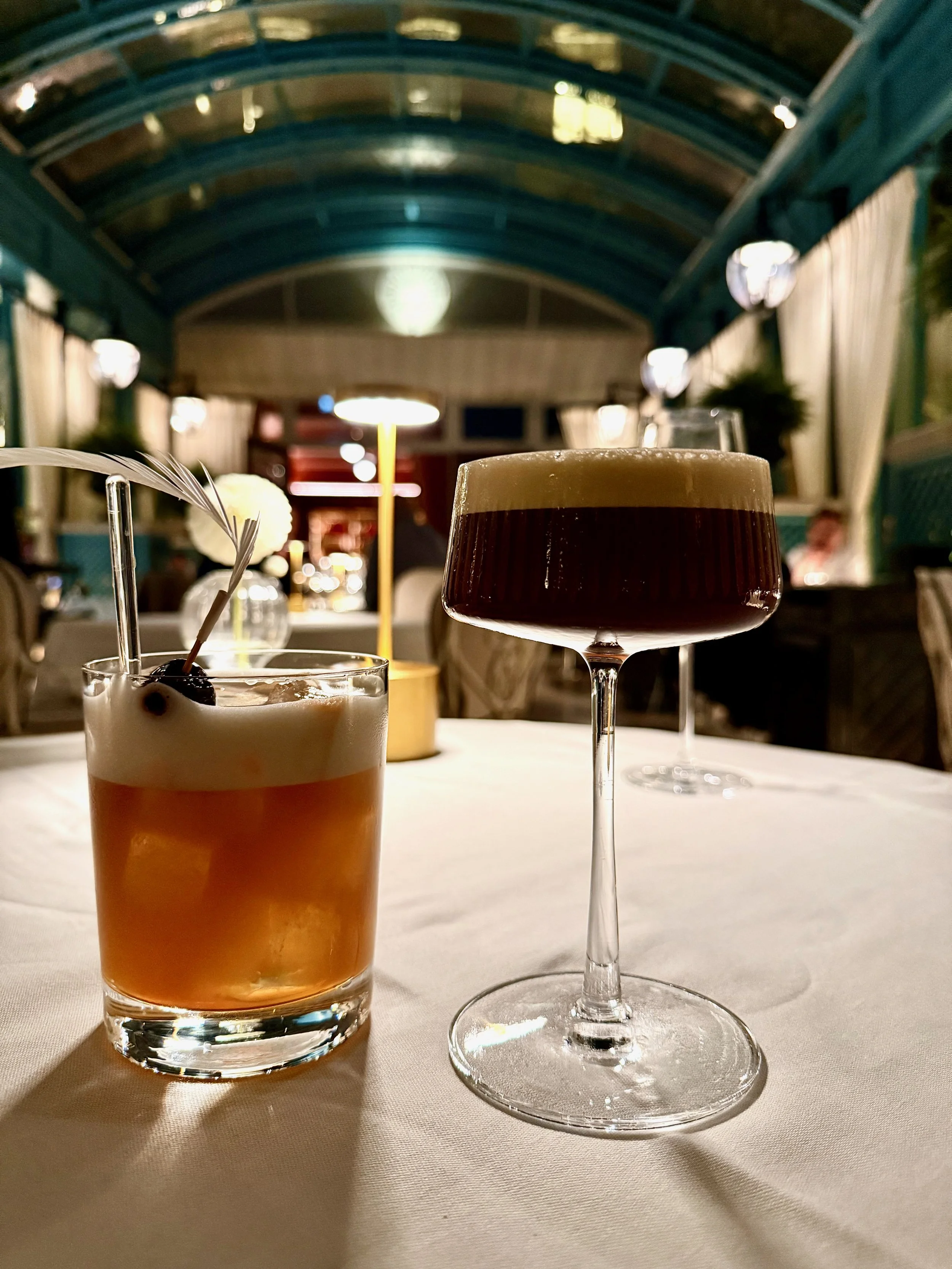 Two cocktails on a white tablecloth in a restaurant with blue arched ceiling, warm lighting, curtains, and floral decor in the background.