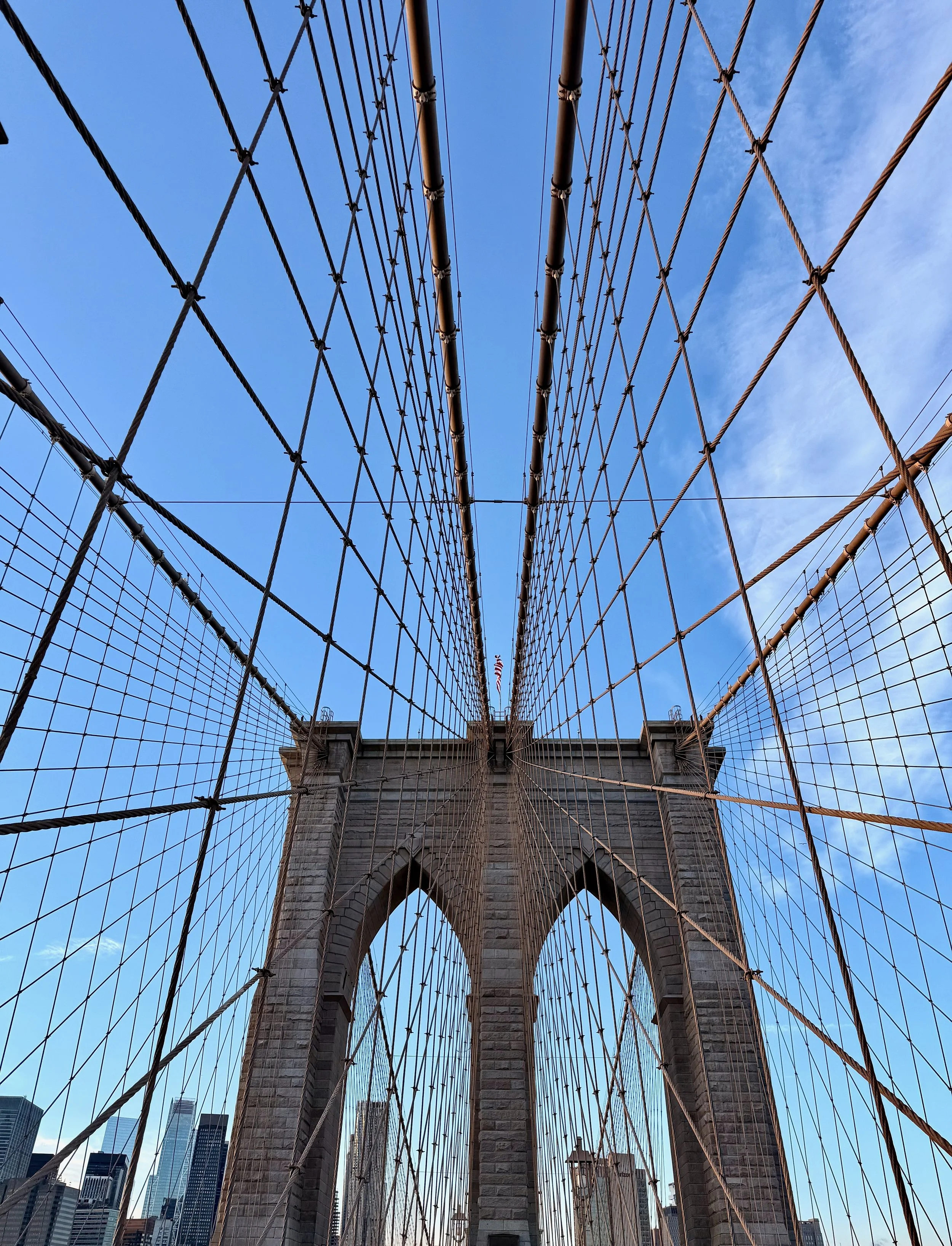 Looking up at the Brooklyn Bridge in New York City, with the stone towers and crisscrossing cables, under a blue sky with a few clouds.