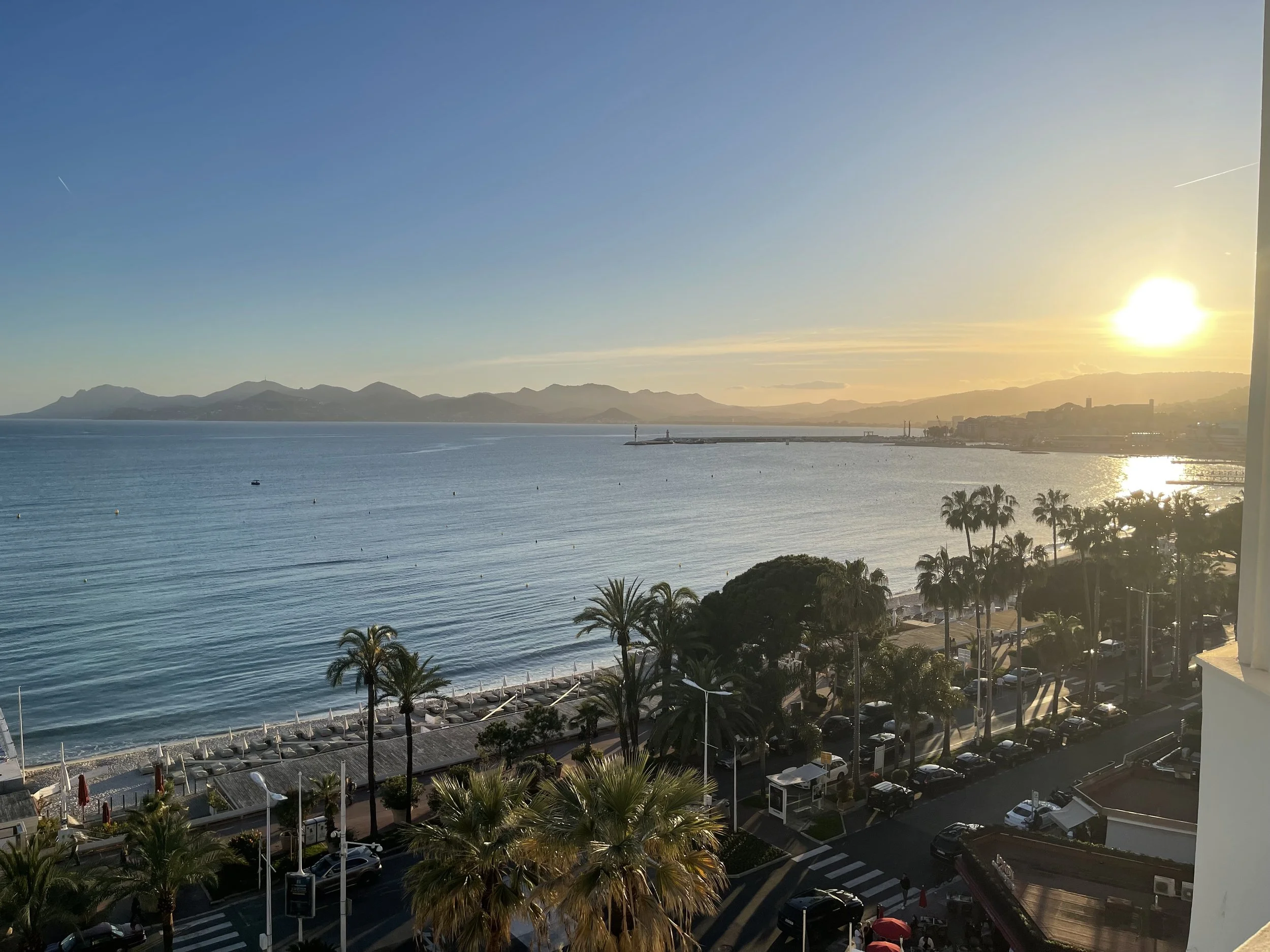A scenic view of a calm ocean at sunset with mountains in the background, palm trees along the coast, and a parking lot in the foreground.