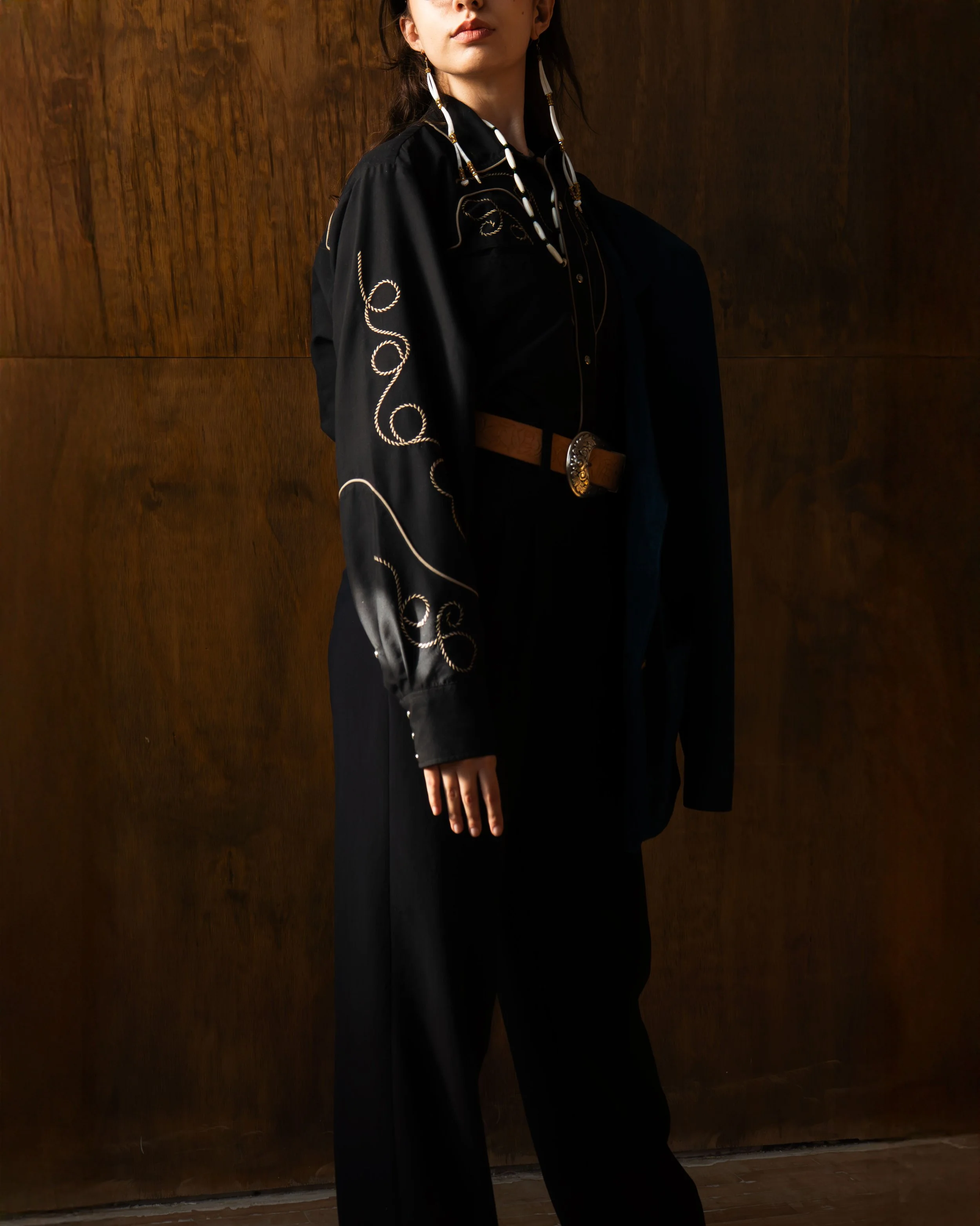 Woman wearing a black blouse with embroidered gold and white designs, black pants, a brown belt, and a necklace with large beads. Standing against a wooden wall.