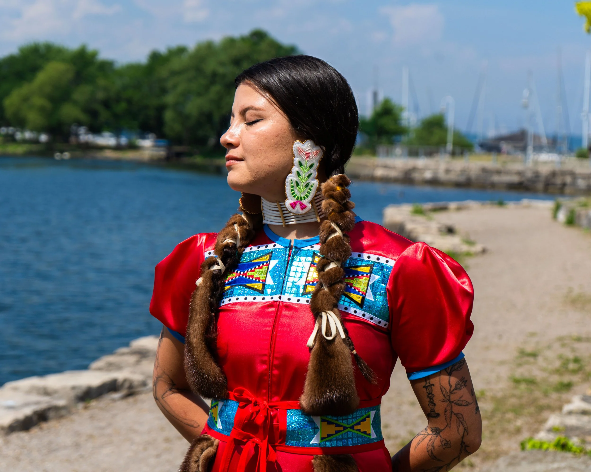 A woman with long braided hair wearing traditional Native American attire, standing outdoors near a body of water with trees and boats in the background.