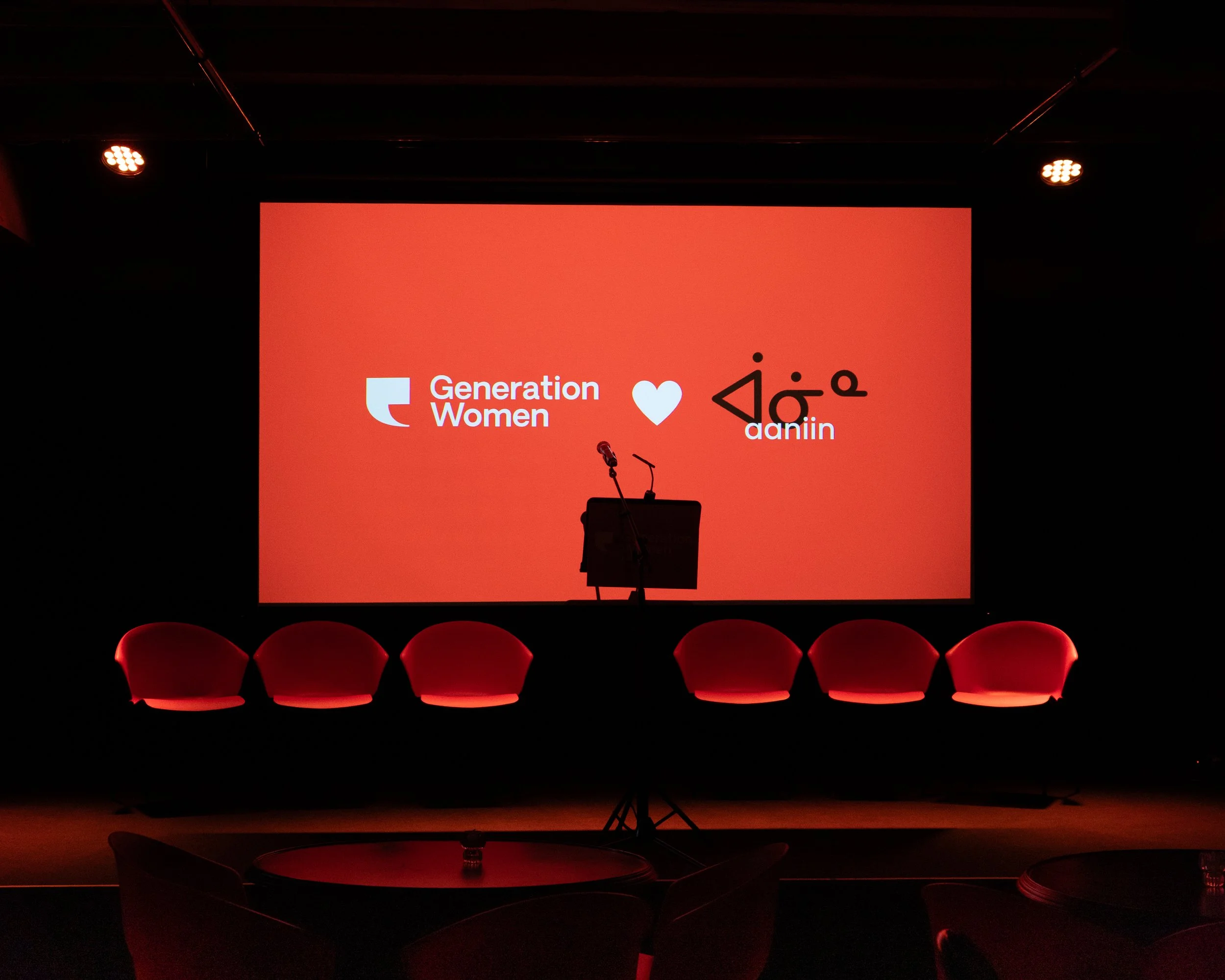 Stage with a large red screen displaying logos for 'Generation Women' and 'Daniin' separated by a heart symbol, five red armchairs in front, microphone and stand centered on stage, dim lighting in the venue.