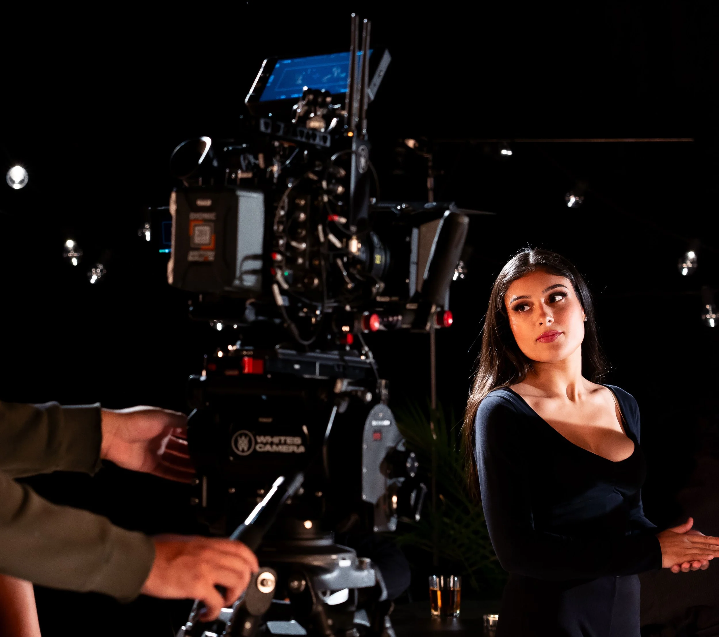 A woman with long dark hair and a black long-sleeve top standing next to professional video camera on a tripod, indoors with black background and dim lighting.