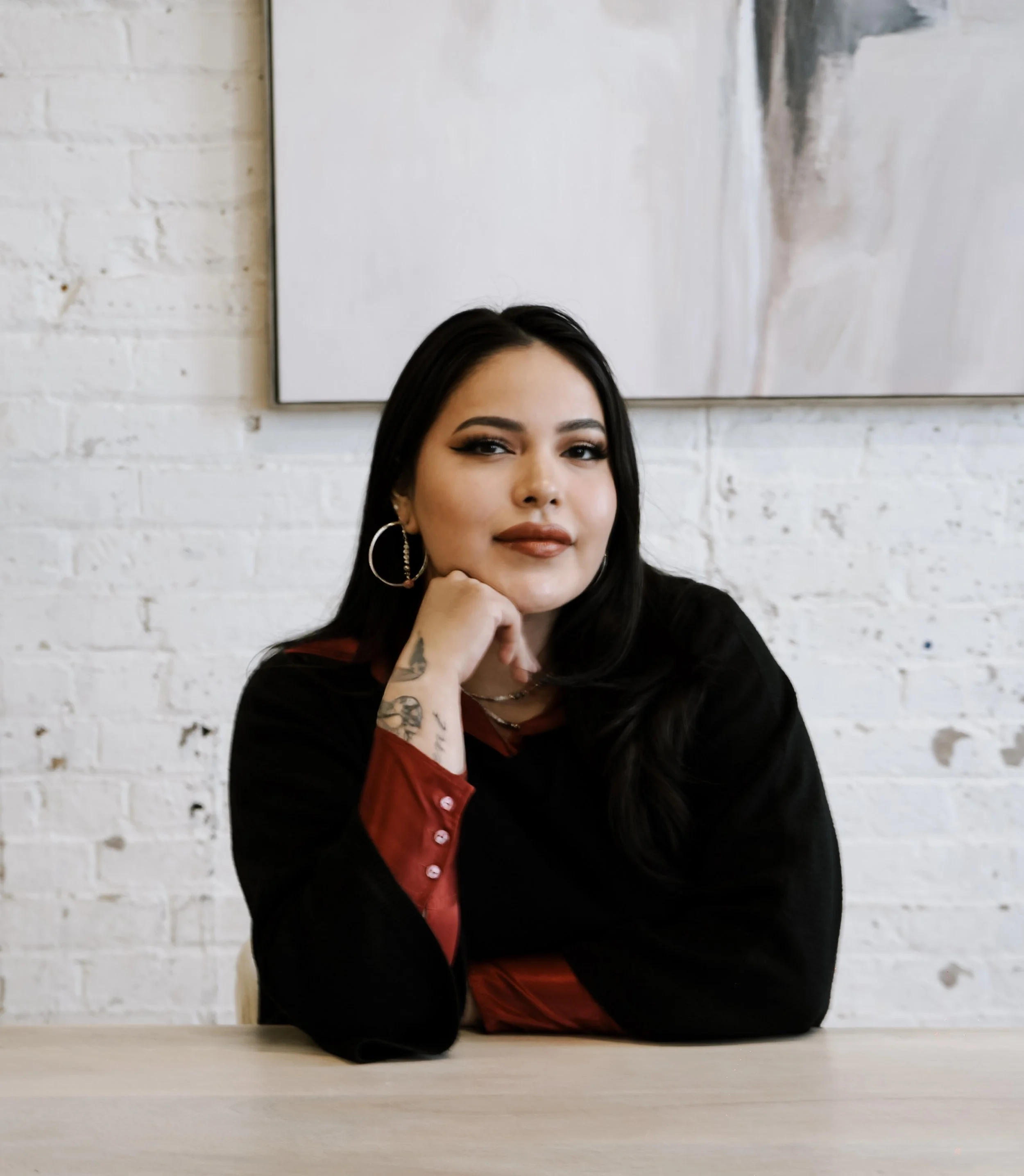 A woman with black hair, wearing earrings, resting her chin on her hand, sitting at a table in front of a white brick wall with abstract artwork.