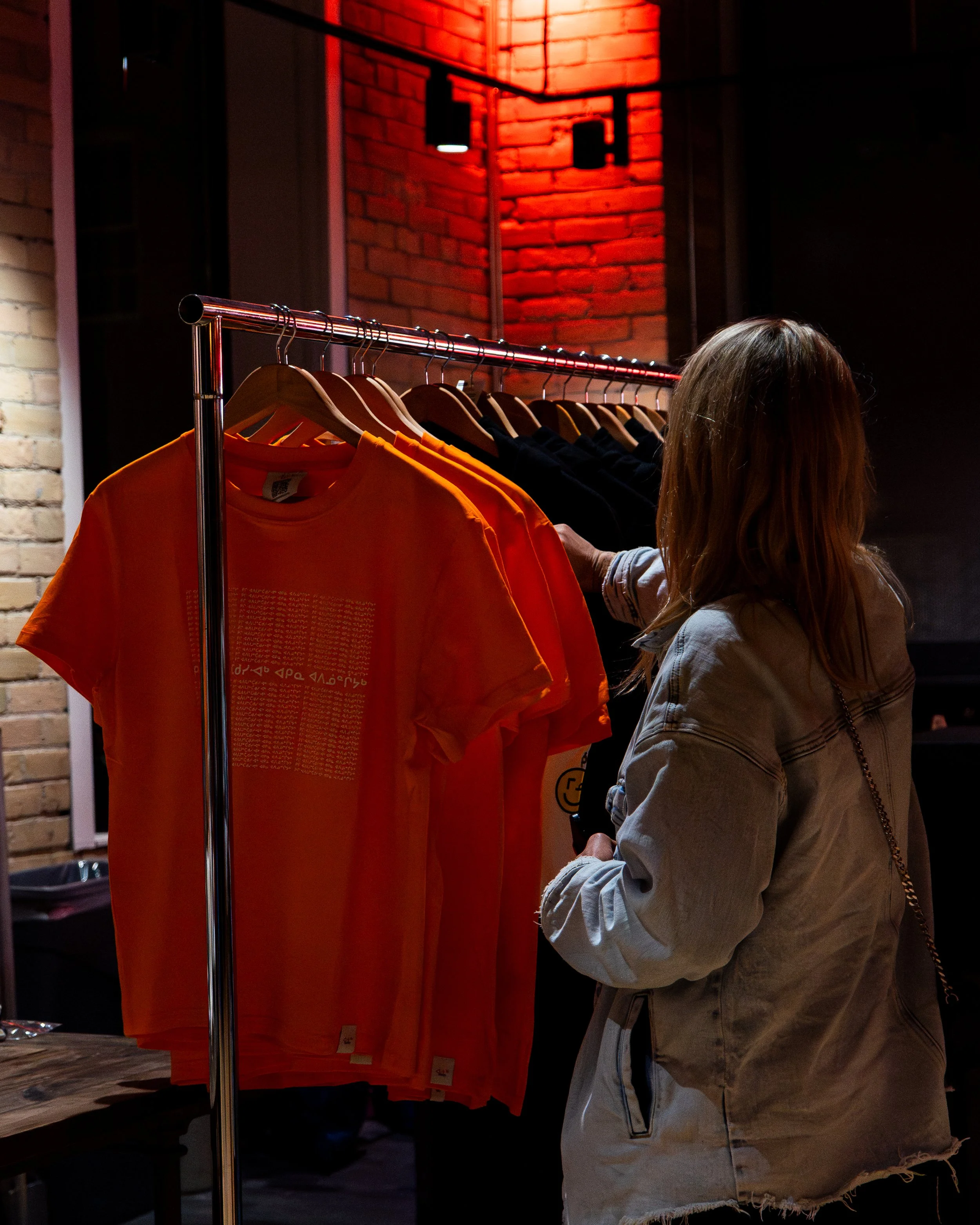 A woman shopping for clothes at a retail store with a rack of orange, black, and other colored shirts, brick wall background, and red lighting.