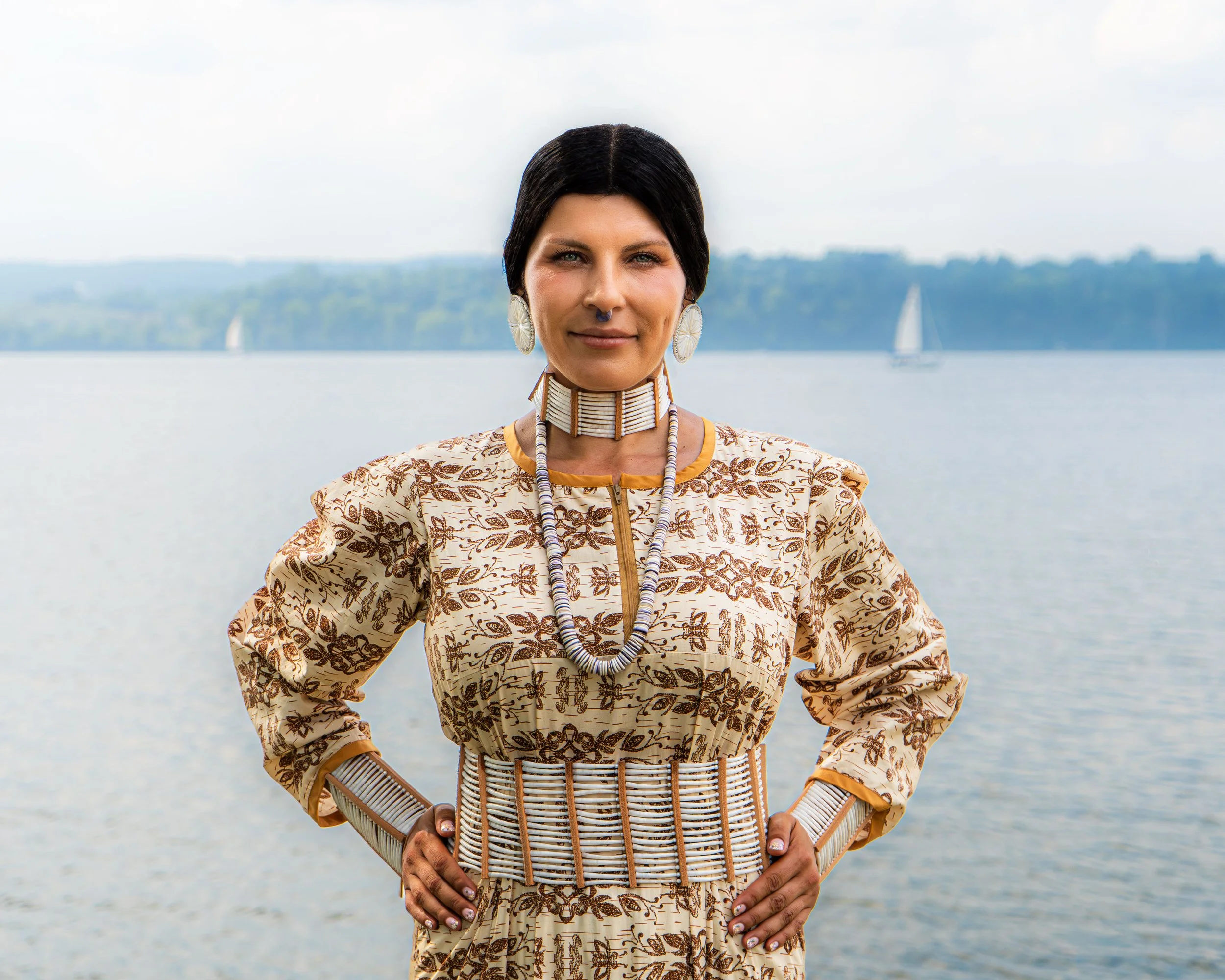 A woman standing outdoors by a body of water, wearing traditional clothing and jewelry, with her hands on her hips, and sailboats visible in the background.