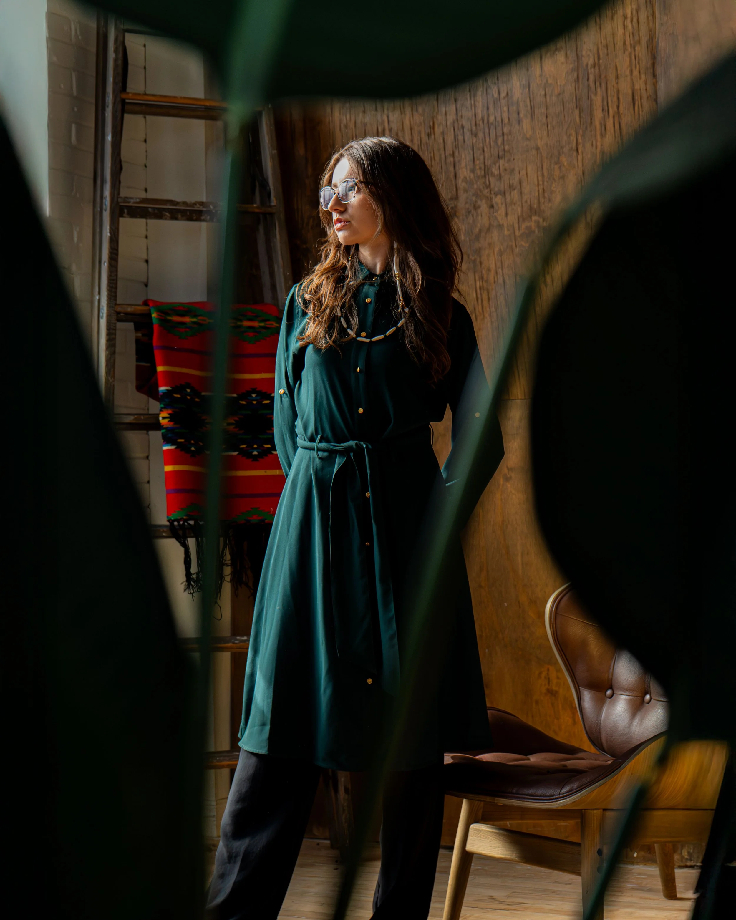 A woman with long brown hair and glasses stands in a room with warm wood walls, wearing a dark green dress and black pants, framed by large green leaves in the foreground.