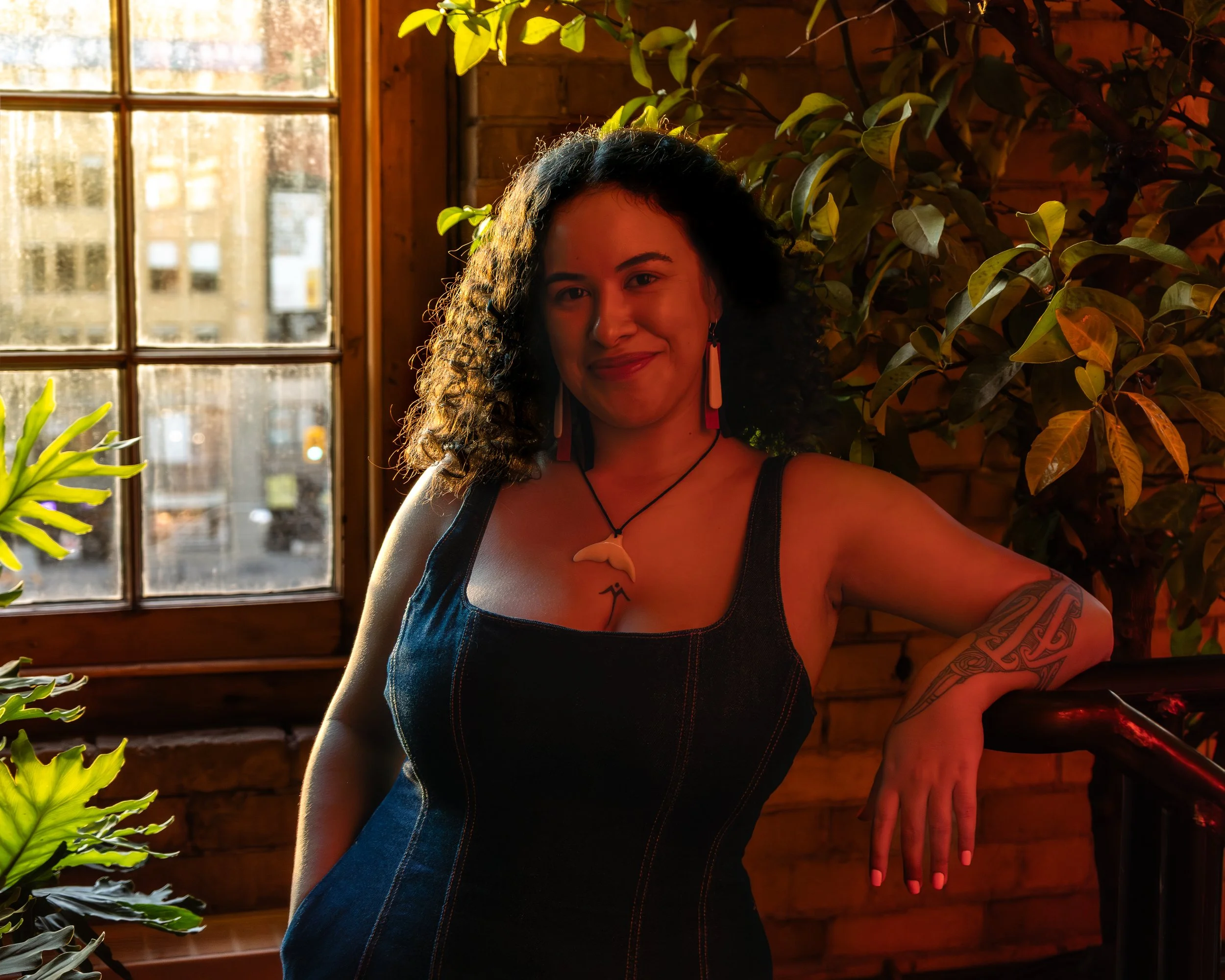 A woman with curly dark hair, wearing a black sleeveless dress, standing indoors beside a window and leafy plants, smiling at the camera.