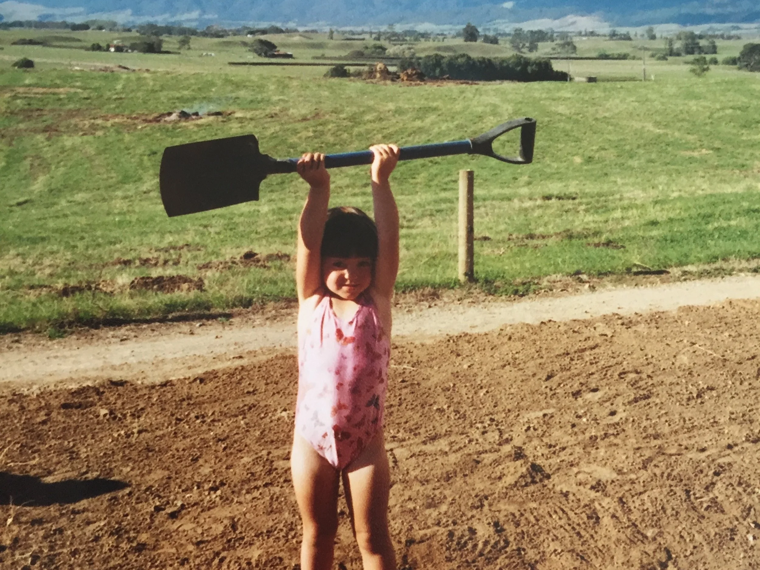 A young girl in a pink swimsuit standing outside on dirt ground, holding a large black shovel above her head with both hands, smiling.