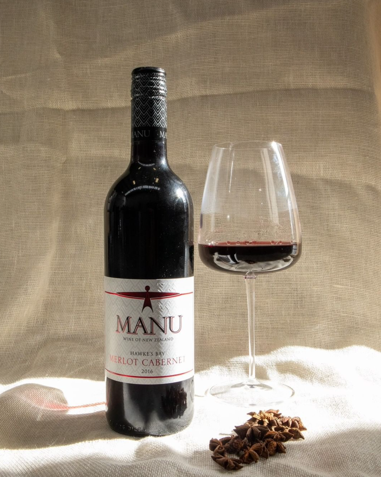 A bottle of Manu Merlot Cabernet wine from New Zealand, a glass of red wine, and a small pile of star anise on a beige fabric background.
