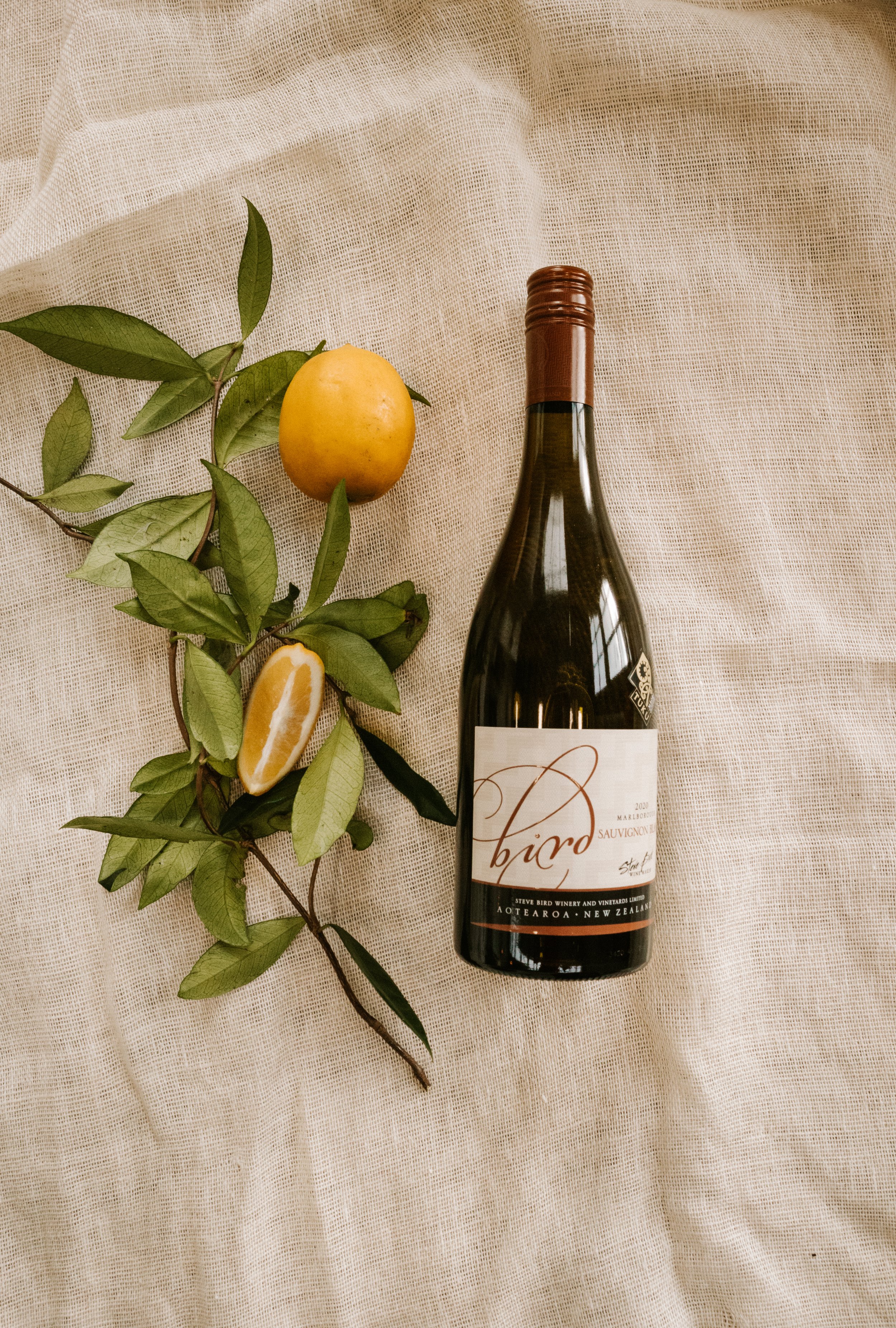 A bottle of red wine, a yellow lemon, and a lemon wedge on a beige fabric background.