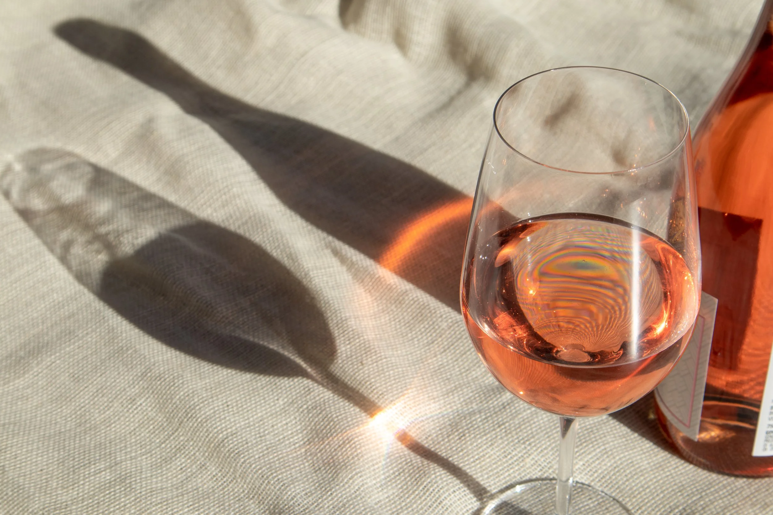 A glass of pink rosé wine casting a shadow and rainbow-like reflections on a beige tablecloth.