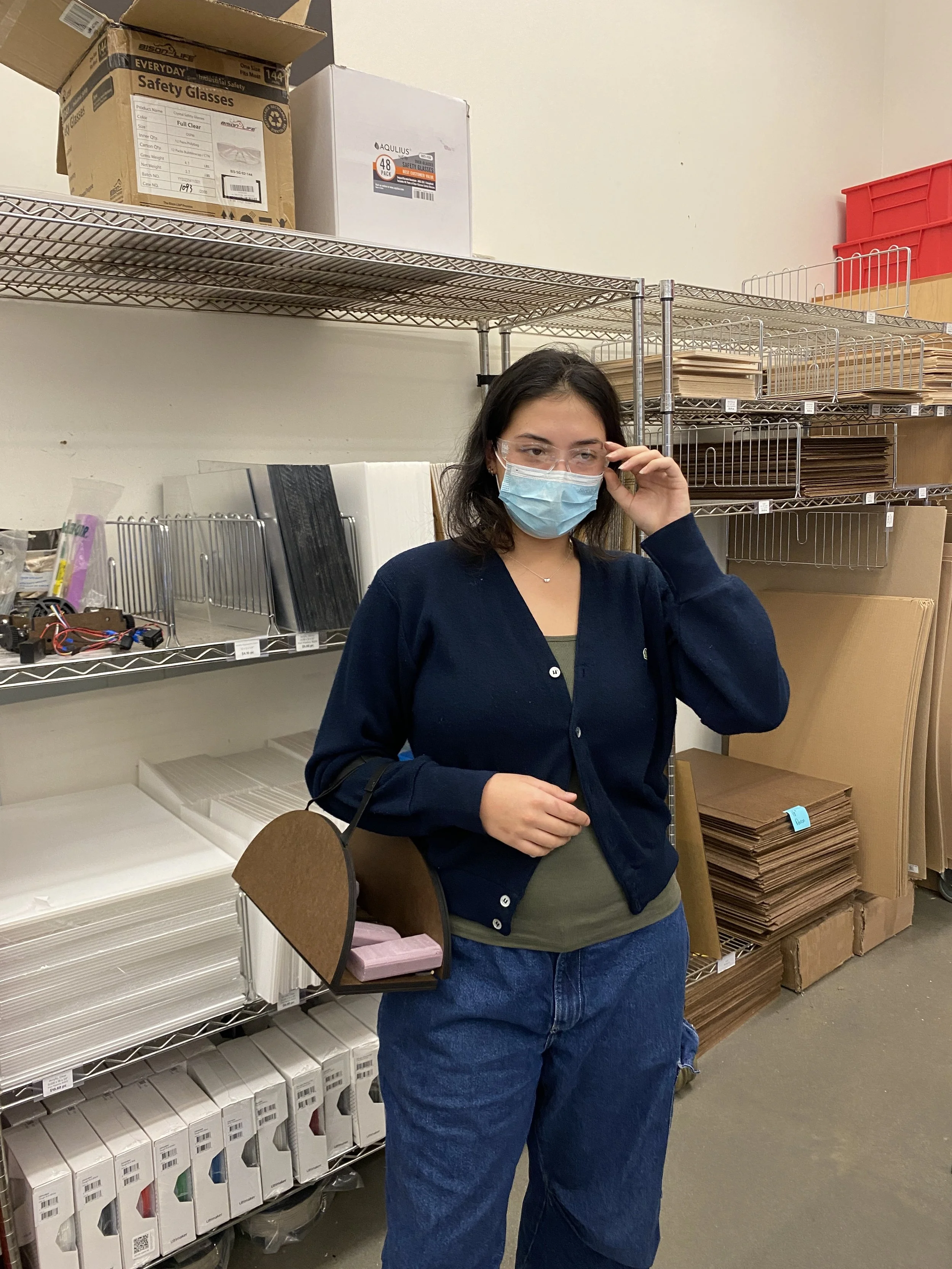 A woman wearing glasses and a face mask adjusting her glasses while standing in a warehouse or storage room with metal shelving units filled with boxes, folders, and office supplies.