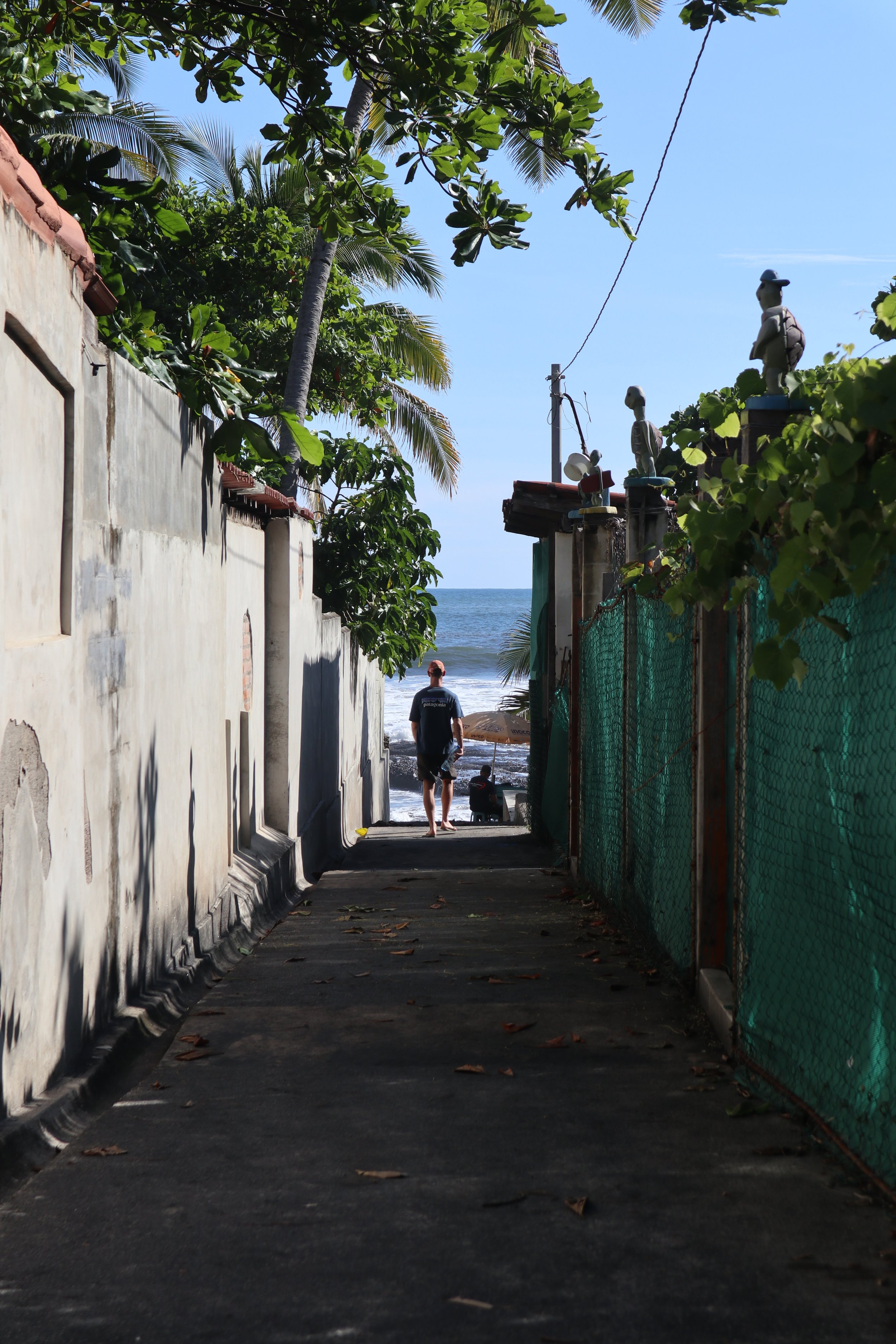 A pathway leading to a beach with a man and a child near the shoreline. The scene includes palm trees, a fence with decorative statues, and a clear blue sky.