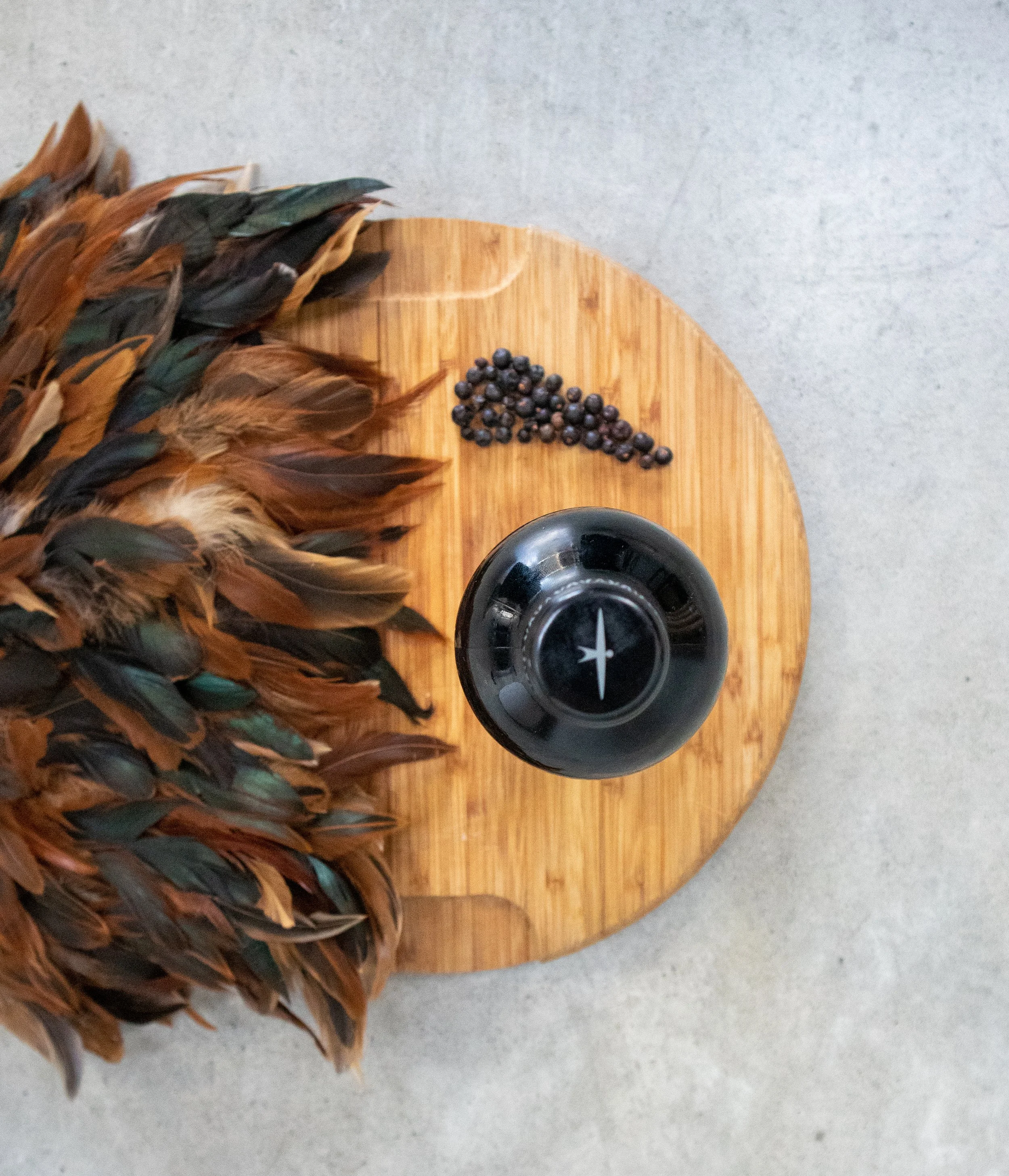 A wooden cutting board with brown feathers on the left, a small pile of black peppercorns, and a black pepper mill against a gray surface.