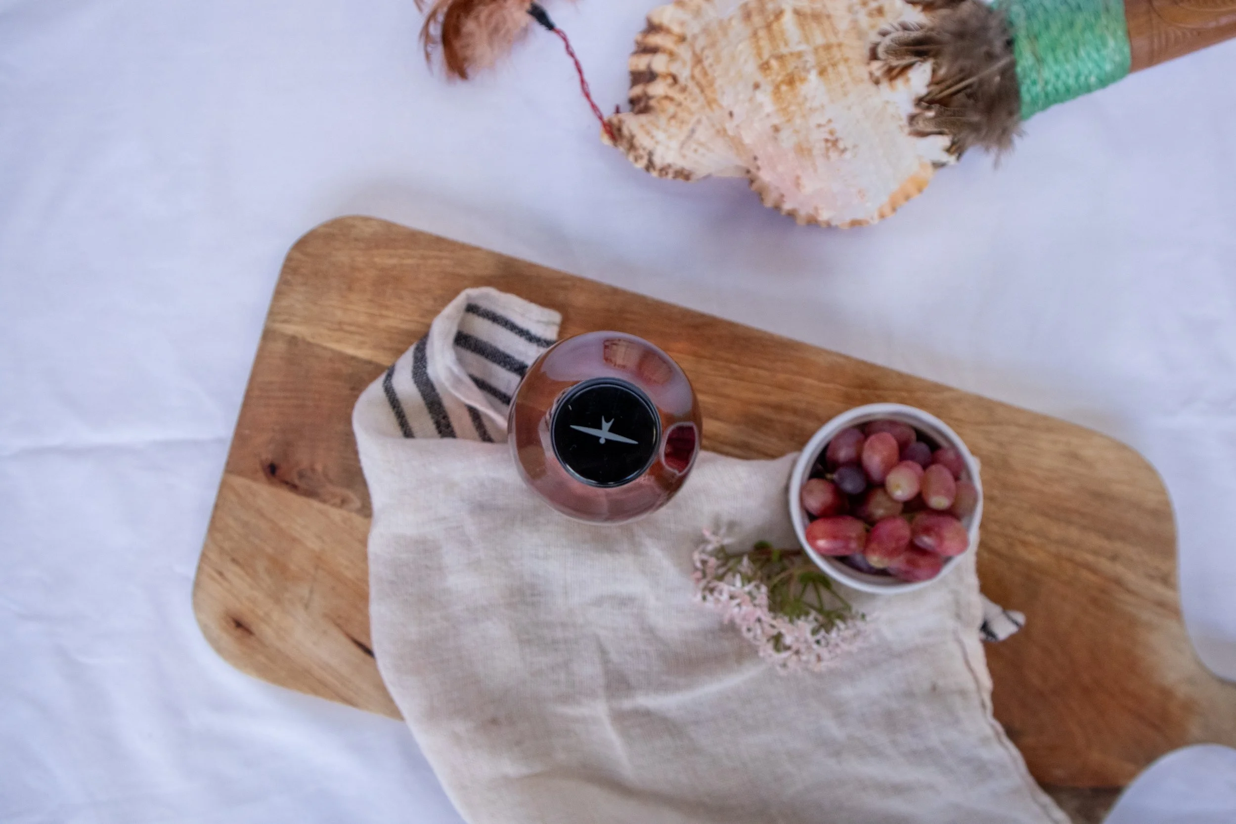 A wooden board with a beige linen cloth, a bowl of grapes, a striped sock, a small vase with pink flowers, and a round decorative object with a shell and feathers on a white tablecloth.