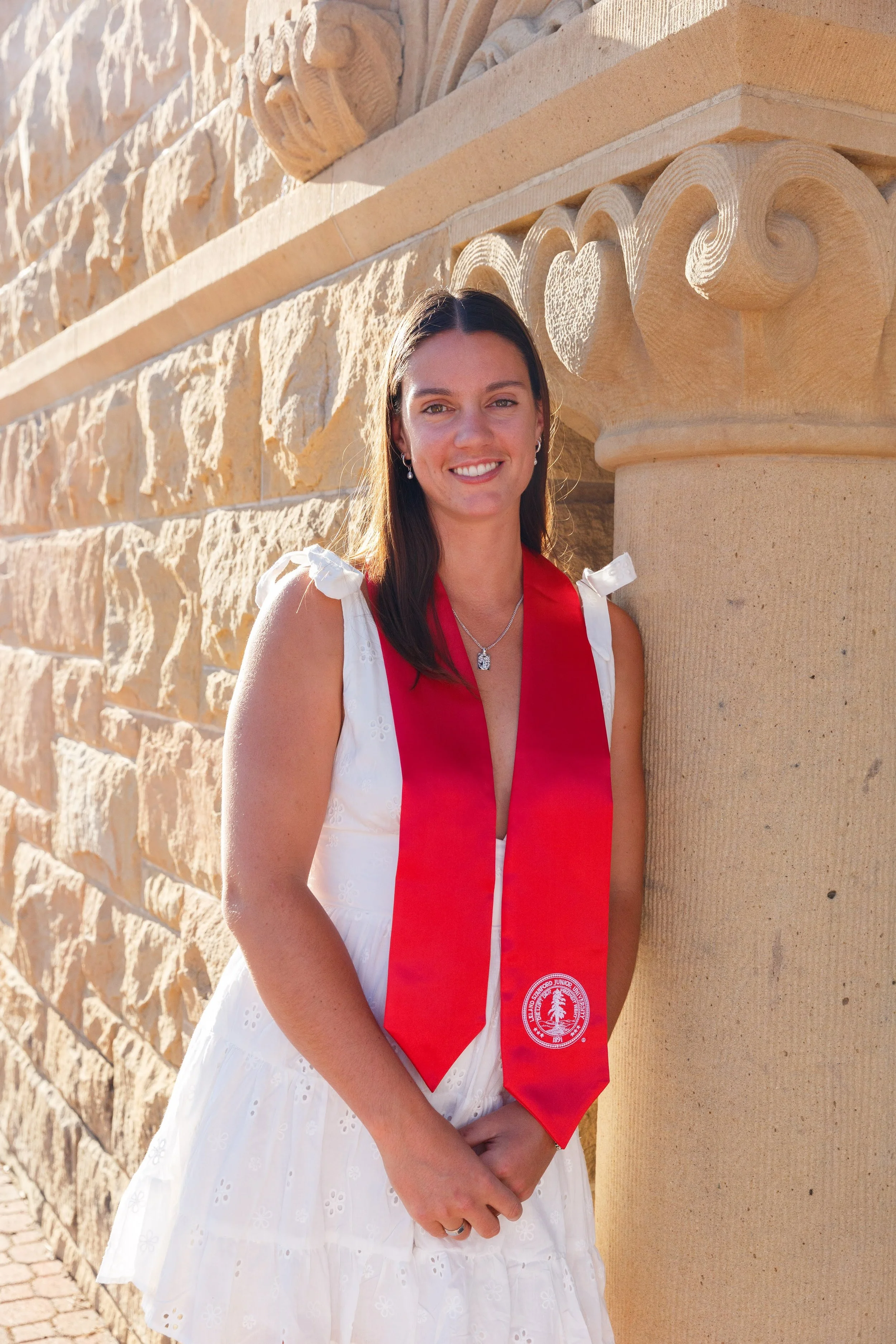 A young woman in a white dress with a red stole, standing outdoors beside a stone wall with intricate carvings, smiling at the camera.