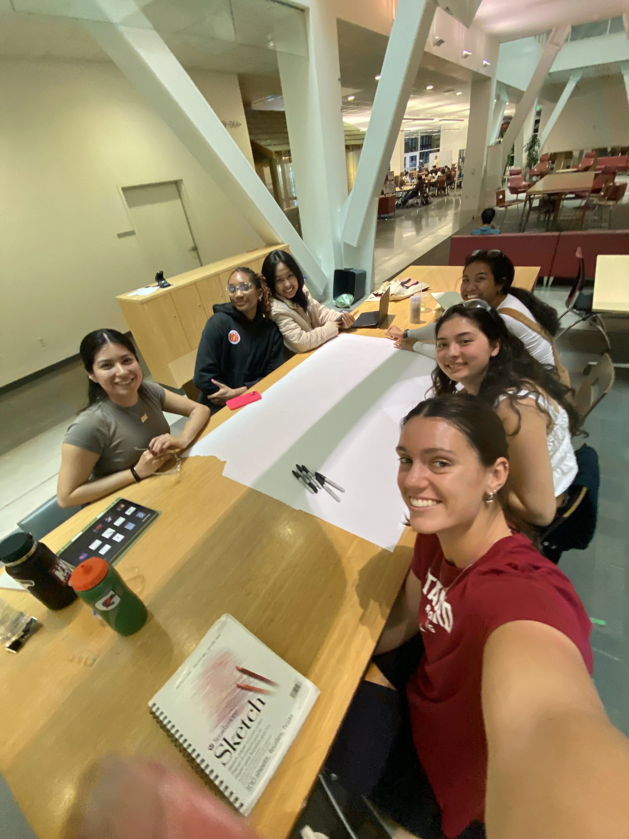 A group of six young women smiling and sitting around a large table in a well-lit, modern indoor space, with notebooks, pens, and drinks on the table.