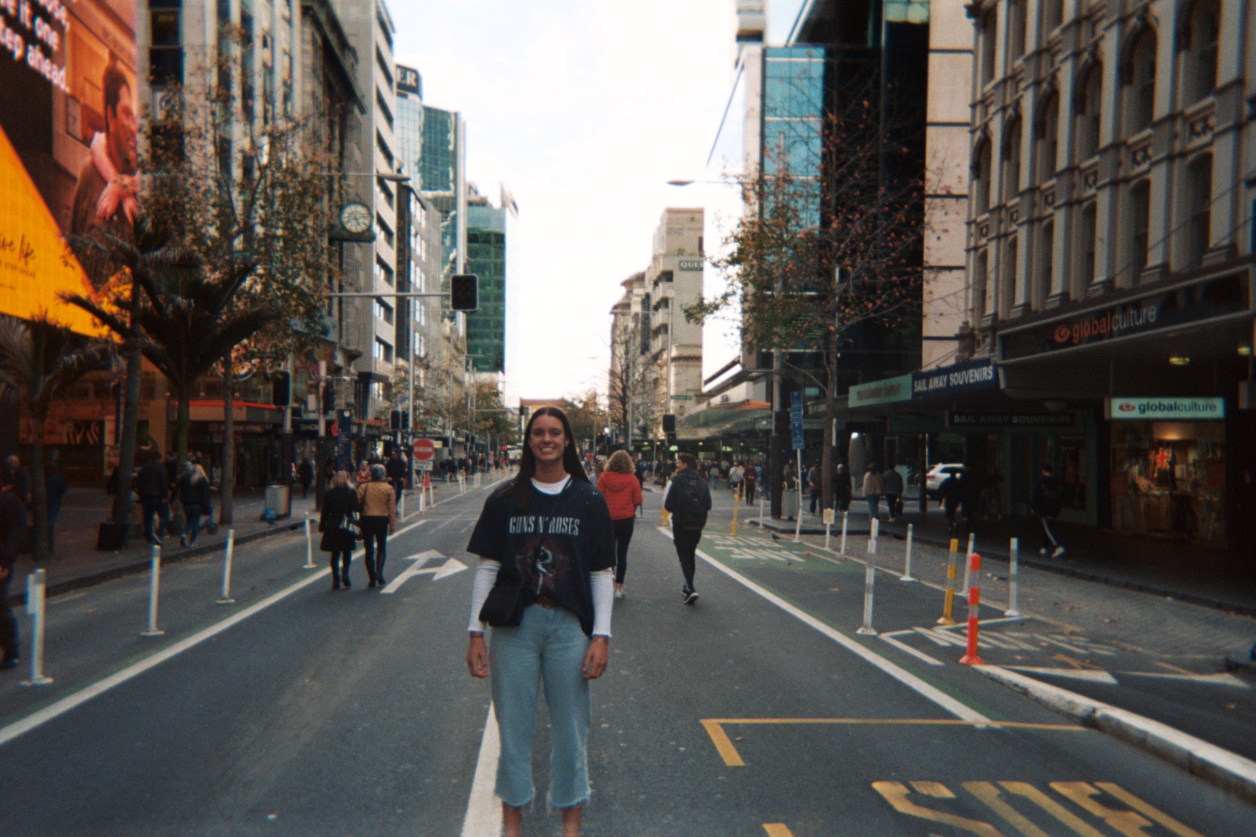 A young woman standing in the middle of a city street with tall buildings on either side, other pedestrians walking around, some trees without leaves, and store signs visible in the background.