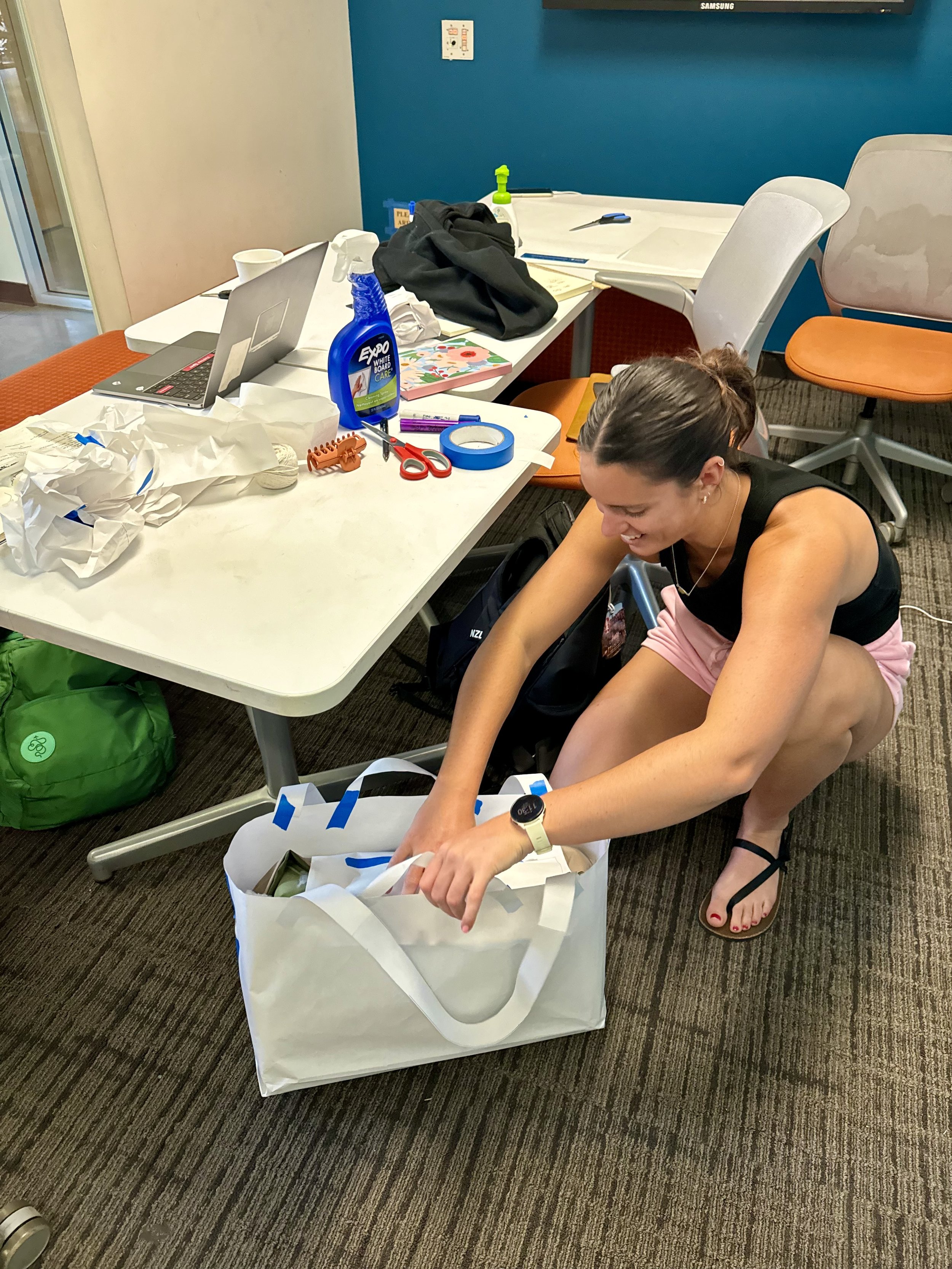 A woman is kneeling on the floor, reaching into a large white shopping bag at her feet. The scene is in a cluttered office or conference room with a table covered in various items, including a laptop, cleaning spray, scissors, tape, and paper. The woman is wearing a black tank top, pink shorts, and sandals.