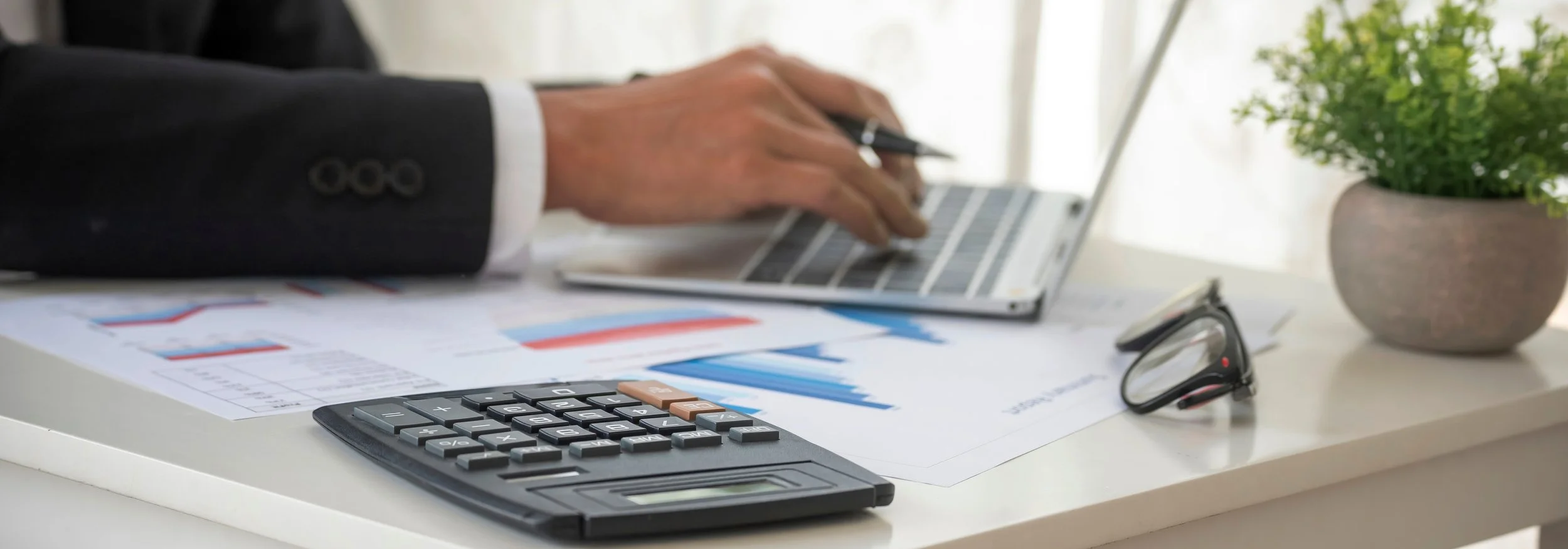 Person in a business suit working at a white desk with a calculator, papers with graphs, a laptop, glasses, and a potted plant.