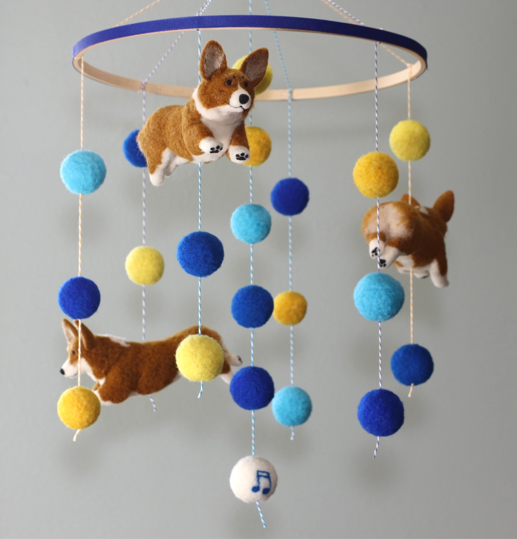 Colorful felt mobile featuring three brown and white felt corgi dogs and multiple felt balls in shades of blue, yellow, and white, hanging from a circular wooden ring.