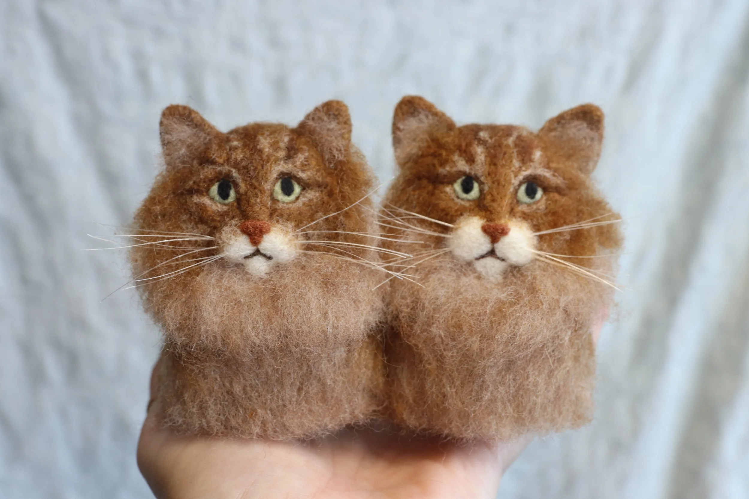 Two handmade felt cat figurines with detailed fur, green eyes, and white and orange facial markings, held in a person's hand against a white background.